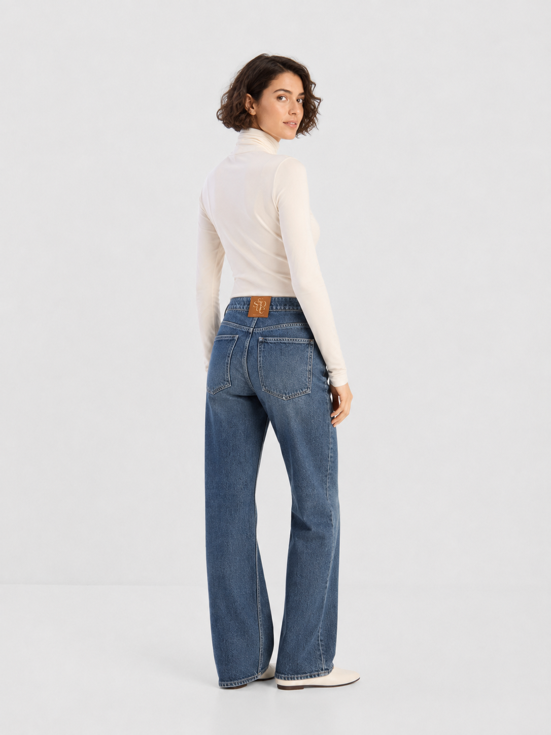 Eine Person mit kurzen Locken trägt ein weißes Langarm-Oberteil und die NOLA Jeans Light Blue – Wide Leg Damenjeans und steht mit dem Rücken zur Kamera, den Kopf über die Schulter gewandt, vor einem schlichten, hellen Hintergrund.