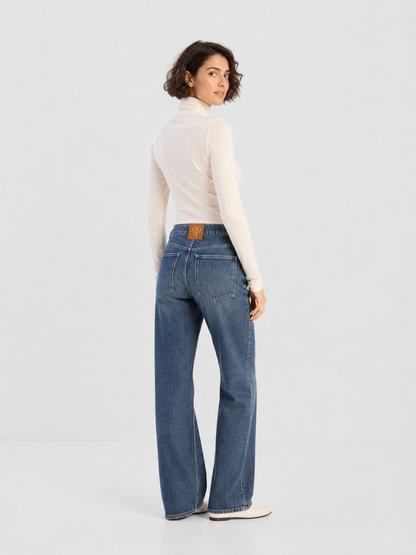 Eine Person mit kurzen Locken trägt ein weißes Langarm-Oberteil und die NOLA Jeans Light Blue – Wide Leg Damenjeans und steht mit dem Rücken zur Kamera, den Kopf über die Schulter gewandt, vor einem schlichten, hellen Hintergrund.