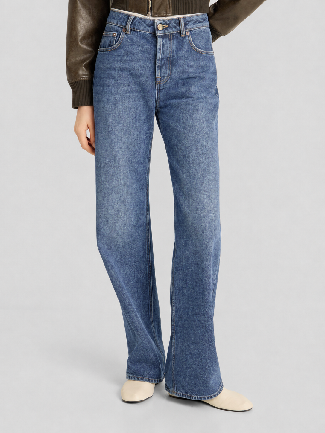 Die Person trägt die NOLA Jeans Light Blue – Wide Leg Damenjeans, kombiniert mit einer braunen Lederjacke und weißen Schuhen, und steht vor einem schlichten grauen Hintergrund. Zu sehen ist nur der Körper von den Schultern abwärts.