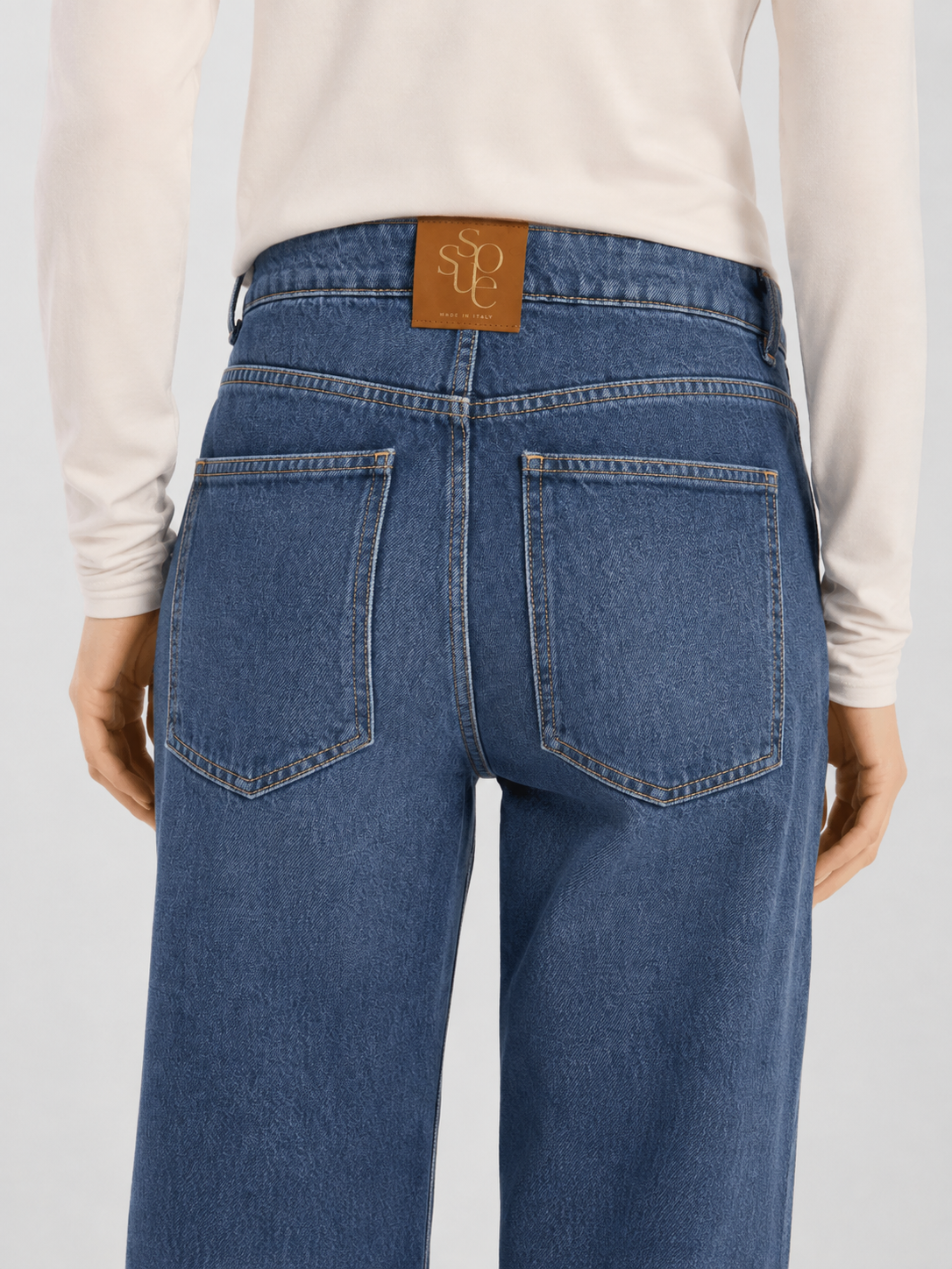Eine Person, die eine hoch taillierte, weit geschnittene NOLA Jeans Light Blue – Wide Leg Damenjeans und ein weißes Langarmshirt trägt, von hinten gesehen. Die Jeans hat zwei Gesäßtaschen und einen braunen Lederaufnäher am Bund.