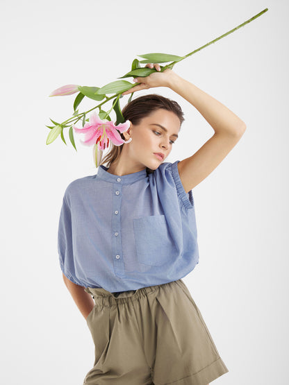 Eine Frau, die eine Cowboy Button Cotton Denim Bluse und olivgrüne Shorts trägt, posiert mit einer rosa Lilienblüte über ihrem Kopf vor einem schlichten weißen Hintergrund.