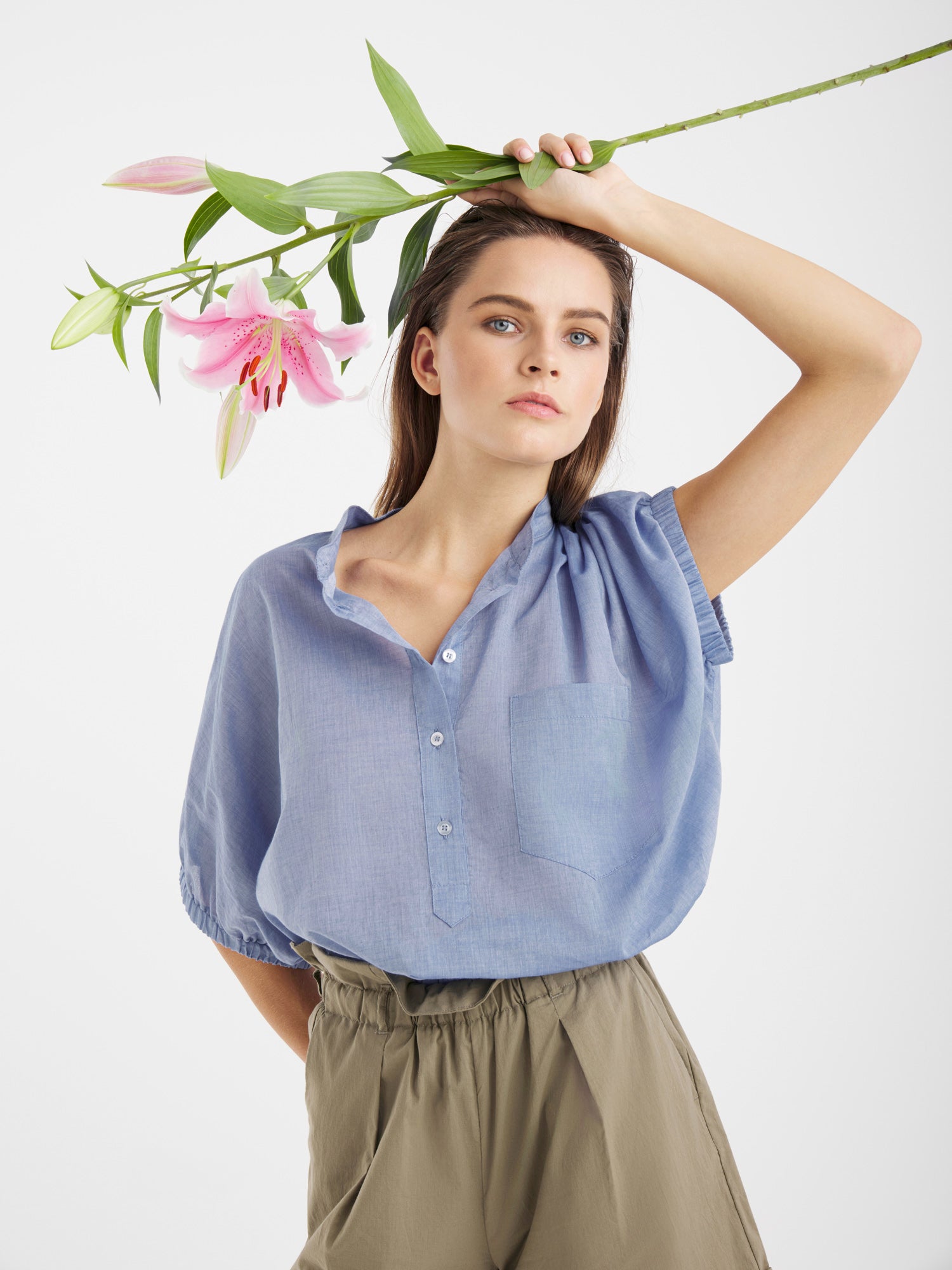 Eine Frau in einem hellblauen Cowboy Button Cotton Denim Hemd hält eine rosa Lilie mit grünen Blättern über ihrem Kopf vor einem schlichten weißen Hintergrund.