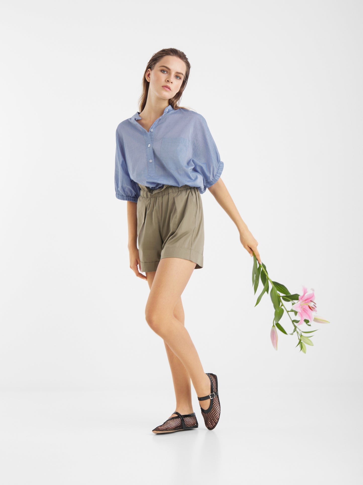 Eine Frau in einem hellblauen Cowboy Button Cotton Denim Hemd und olivgrünen Shorts steht auf einem Bein und hält rosa Lilien hinter sich. Sie trägt schwarze Netzschuhe und posiert vor einem schlichten weißen Hintergrund. Hergestellt in Portugal.