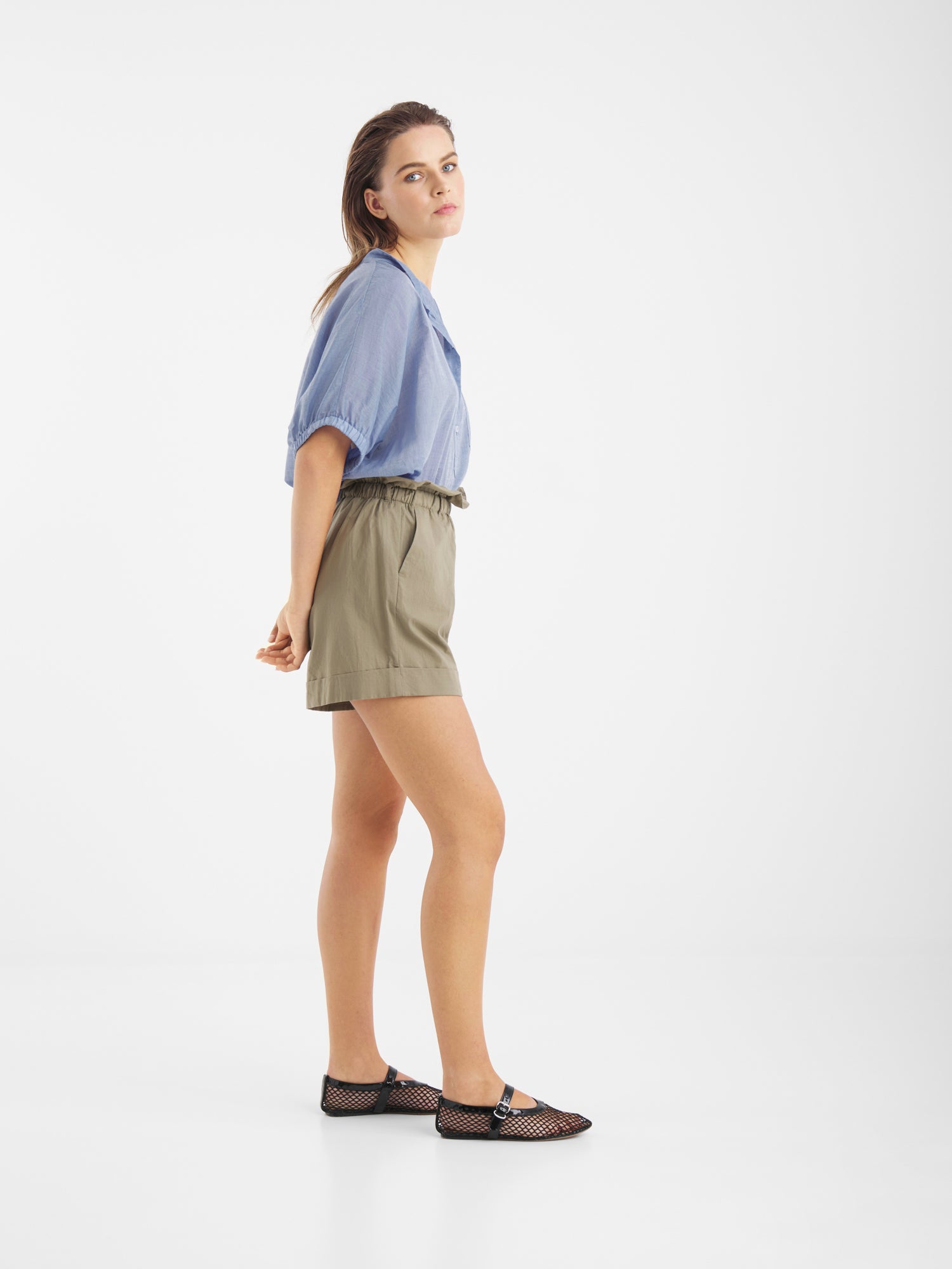 Eine Frau steht seitlich vor einem weißen Hintergrund. Sie trägt ein hellblaues Cowboy Button Cotton Denim Hemd, khakifarbene Shorts mit hoher Taille und schwarze flache Schuhe mit Mesh-Einsätzen. Sie wirft einen Blick über ihre Schulter. Hergestellt in Portugal.