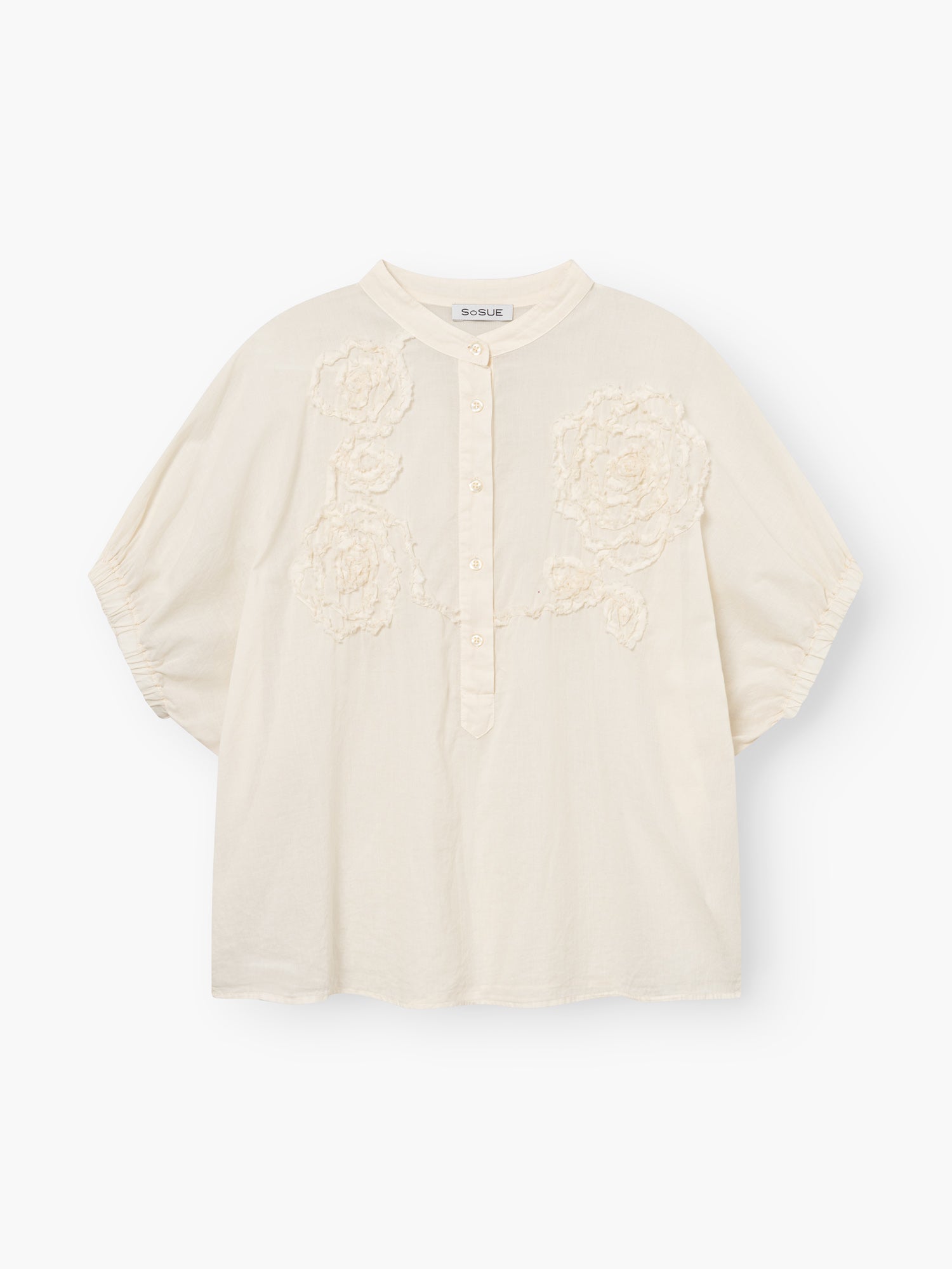 Die Cowboy Crafted Panna ist eine locker sitzende, cremefarbene Bluse mit kurzen Ärmeln, einem Rundkragen, einer teilweisen Knopfleiste und einer strukturierten Blumenstickerei auf der Vorderseite.
