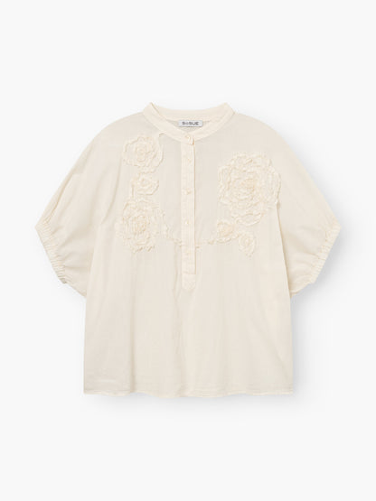 Die Cowboy Crafted Panna ist eine locker sitzende, cremefarbene Bluse mit kurzen Ärmeln, einem Rundkragen, einer teilweisen Knopfleiste und einer strukturierten Blumenstickerei auf der Vorderseite.