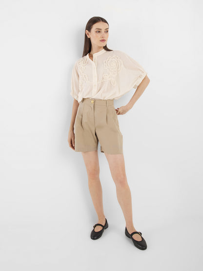 Eine Frau mit glattem braunem Haar und neutralem Gesichtsausdruck trägt die Cowboy Crafted Panna: eine lockere, kurzärmelige cremefarbene Bluse mit Stickerei, beigefarbene Shorts mit hoher Taille und schwarze Mary-Jane-Schuhe, die vor einem weißen Hintergrund stehen.