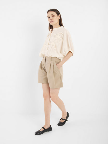 Eine Frau mit glattem braunem Haar steht vor einem weißen Hintergrund und trägt Cowboy Crafted Panna - eine cremefarbene bestickte Bluse, beigefarbene Shorts mit hoher Taille und schwarze Mary-Jane-Schuhe - und eine Hand in ihrer Tasche.