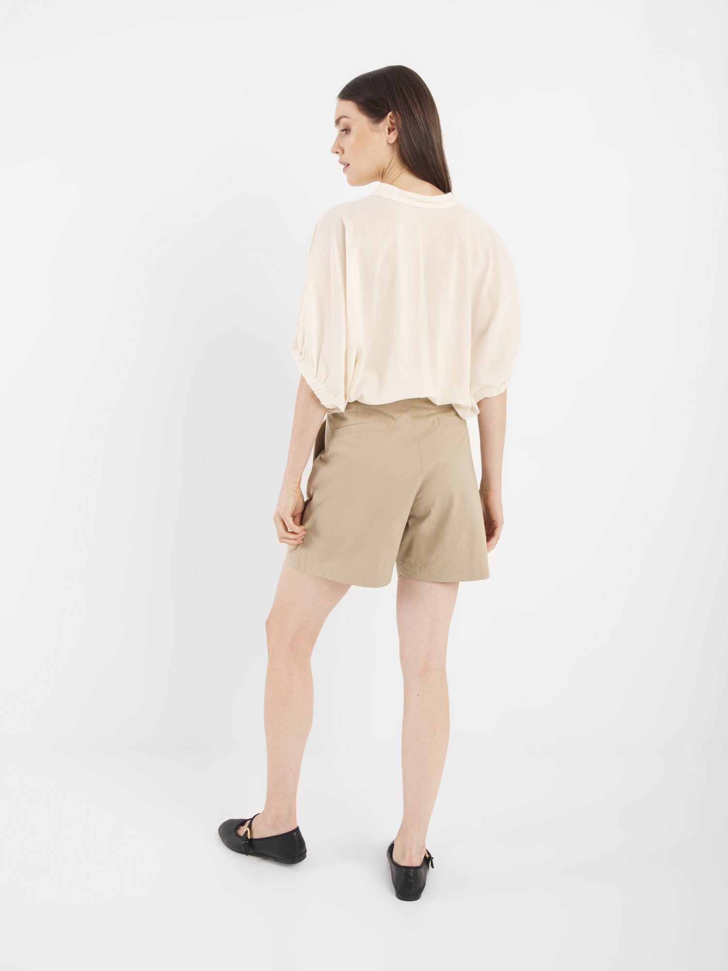 Eine Frau blickt von der Kamera weg und trägt Cowboy Crafted Panna - eine lockere cremefarbene Bluse, hellbraune Shorts und schwarze Flats - vor einem schlichten weißen Hintergrund.