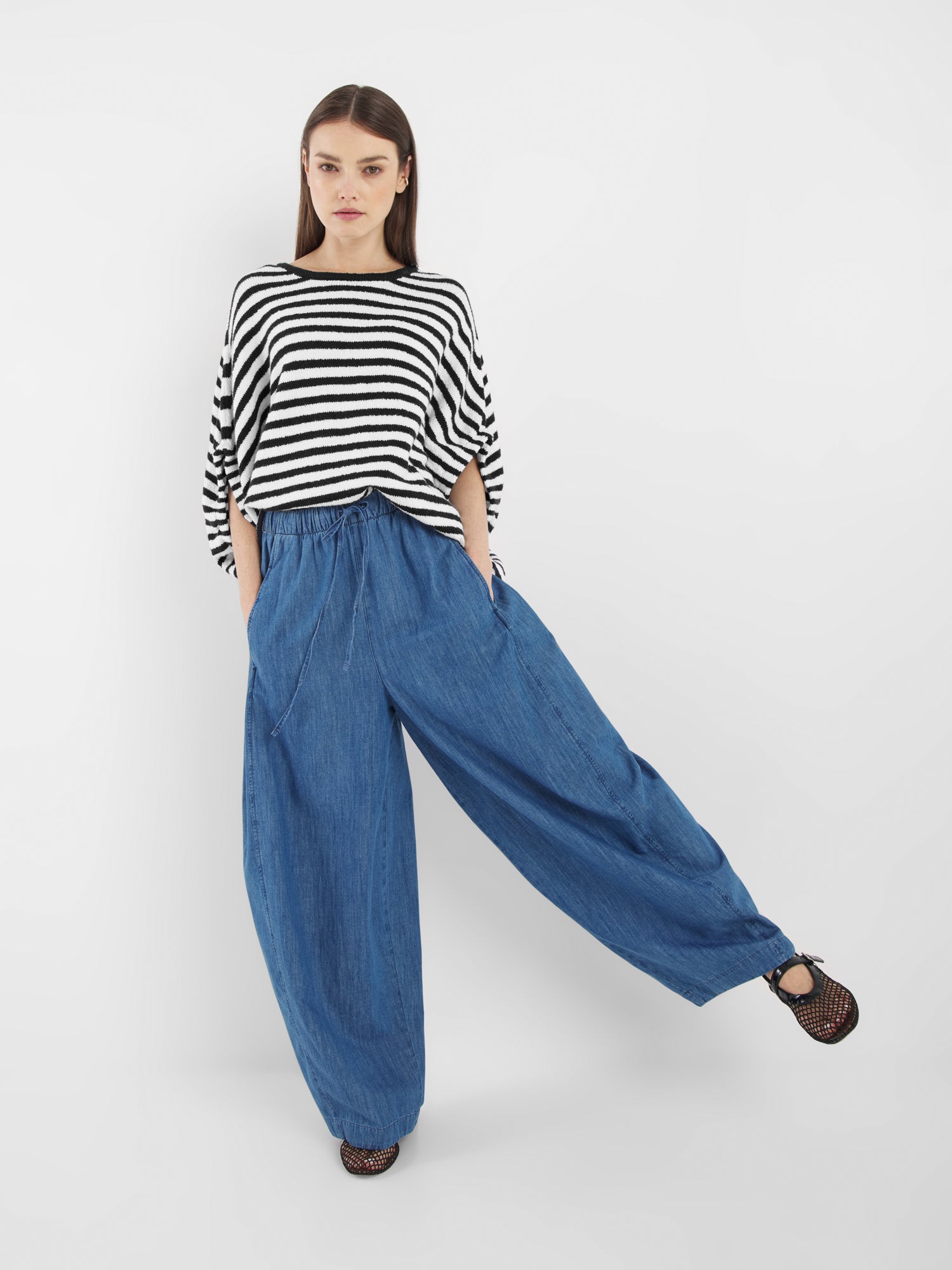 Eine Frau steht selbstbewusst vor einem weißen Hintergrund und trägt die weite blaue Hose von Lizzy Denim mit einem schwarz-weiß gestreiften Oberteil und schwarzen Schuhen. Ein Bein ist angehoben, die Hände in den Taschen, während sie in die Kamera schaut.