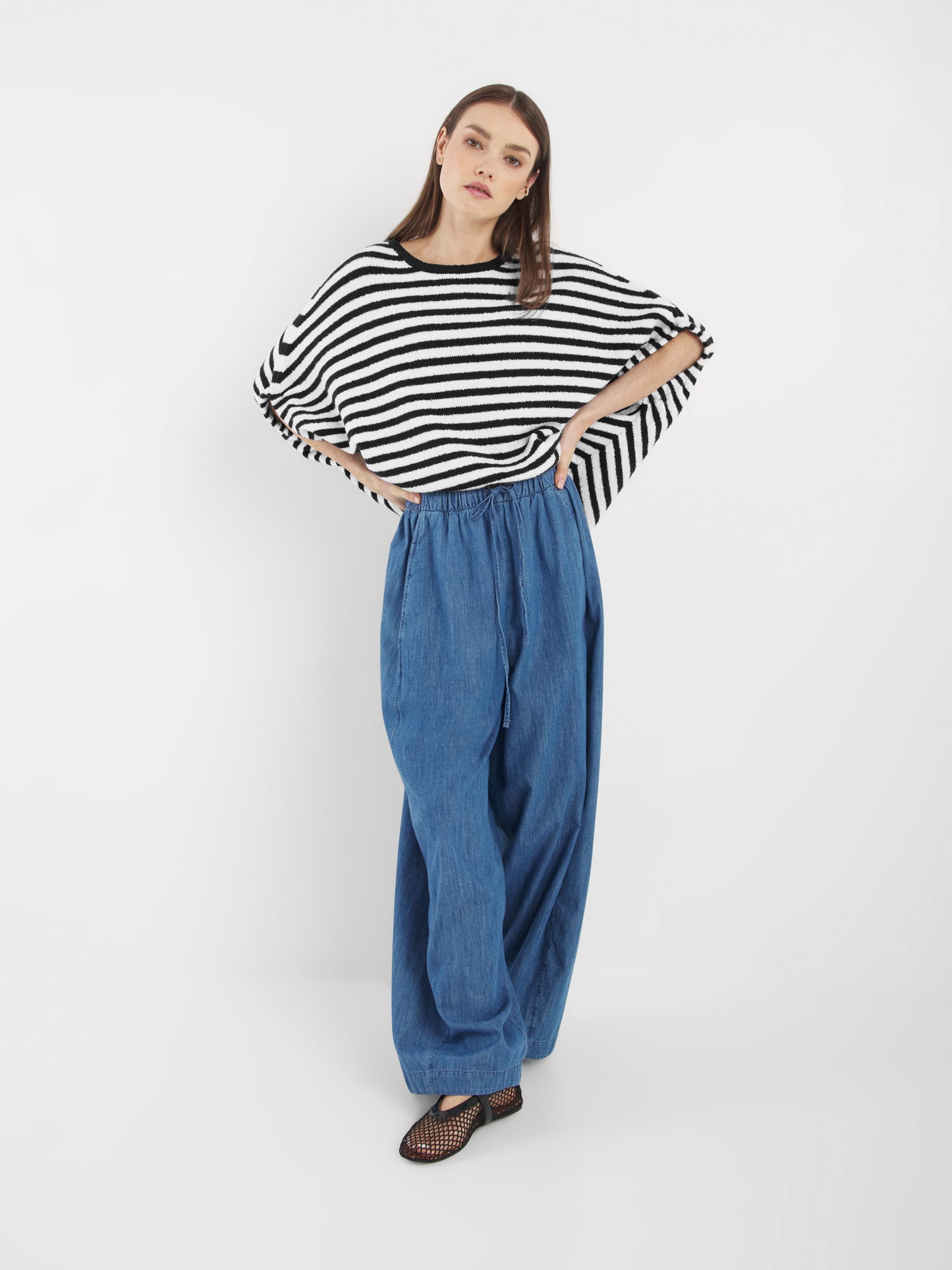 Eine Frau mit glattem braunem Haar und neutralem Gesichtsausdruck trägt das Oberteil Cowboy Knit Stripes Blue White im Poncho-Stil, eine weite blaue Hose mit Kordelzug und schwarze Netzschuhe vor einem weißen Hintergrund.