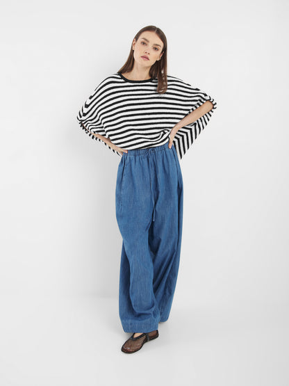 Eine Frau mit glattem braunem Haar und neutralem Gesichtsausdruck trägt das Oberteil Cowboy Knit Stripes Blue White im Poncho-Stil, eine weite blaue Hose mit Kordelzug und schwarze Netzschuhe vor einem weißen Hintergrund.