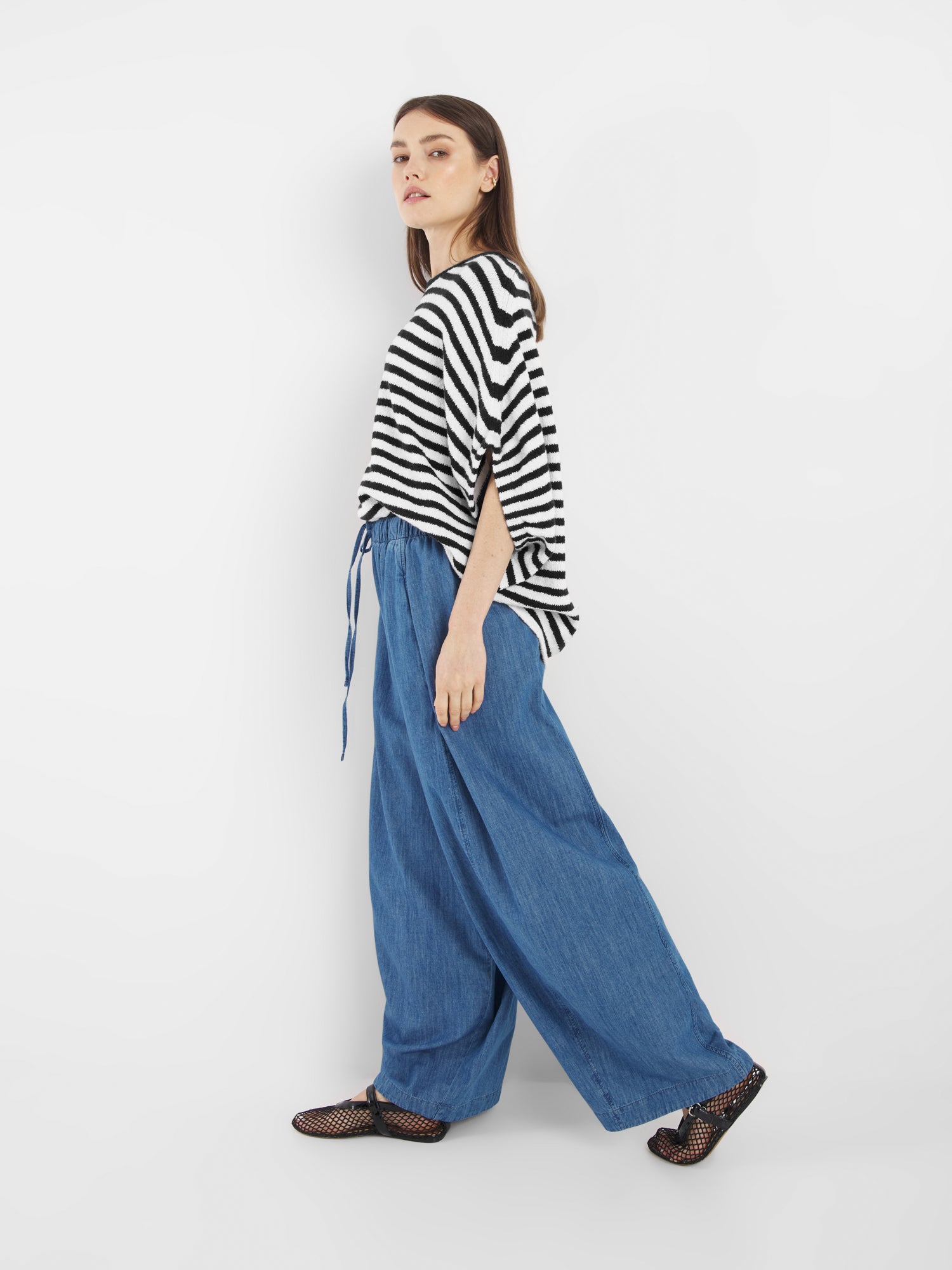 Eine Frau steht seitlich in dem Cowboy Knit Stripes Blue White Oberteil, gepaart mit einer blauen Jeanshose mit weitem Bein und schwarzen Netzschuhen vor einem schlichten weißen Hintergrund.