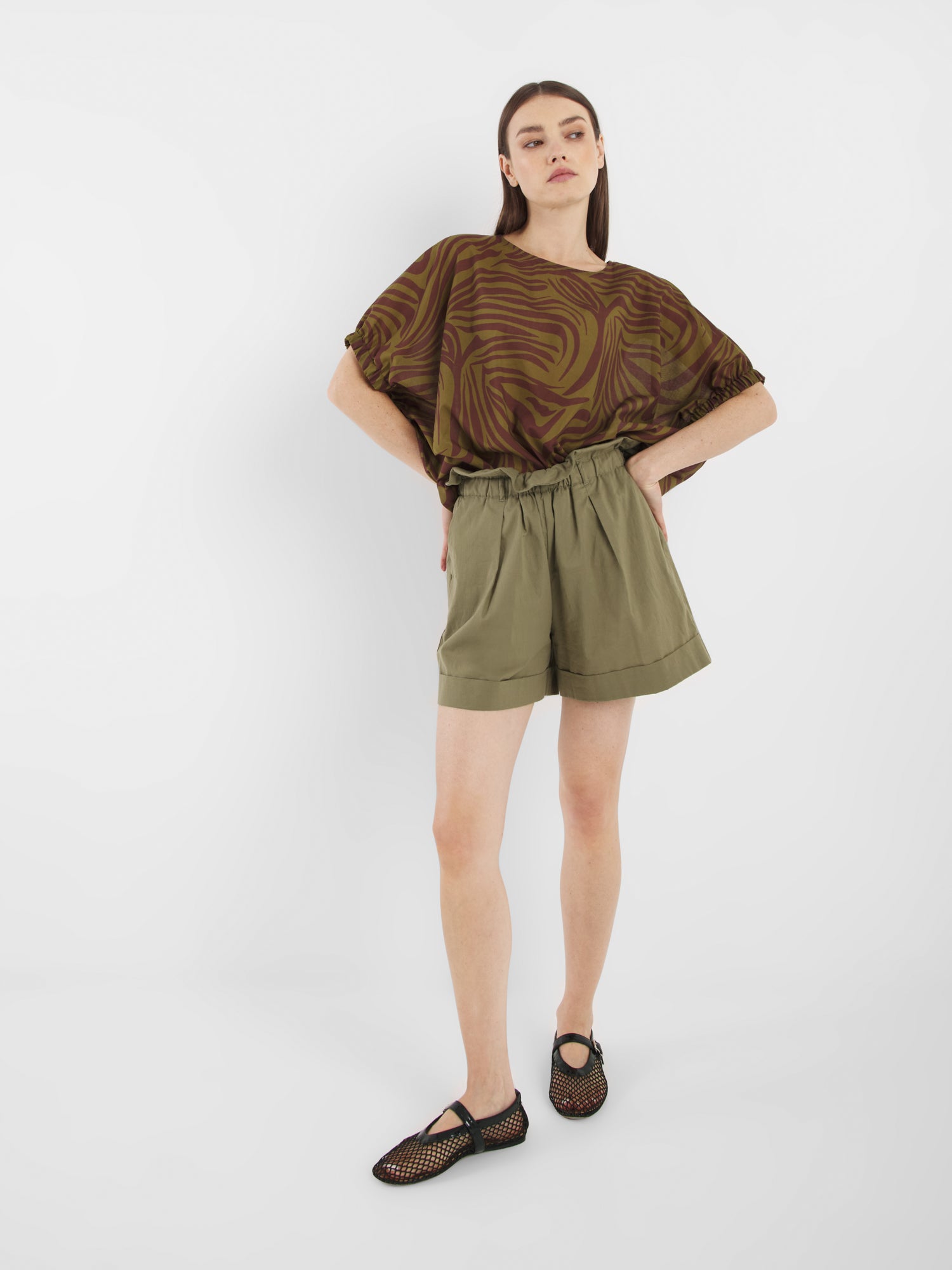 Eine Frau mit glattem braunem Haar und neutralem Gesichtsausdruck steht vor einem weißen Hintergrund und trägt die Bluse Cowboy Zebra Olive, olivgrüne Shorts mit hoher Taille und schwarze gewebte Schuhe.