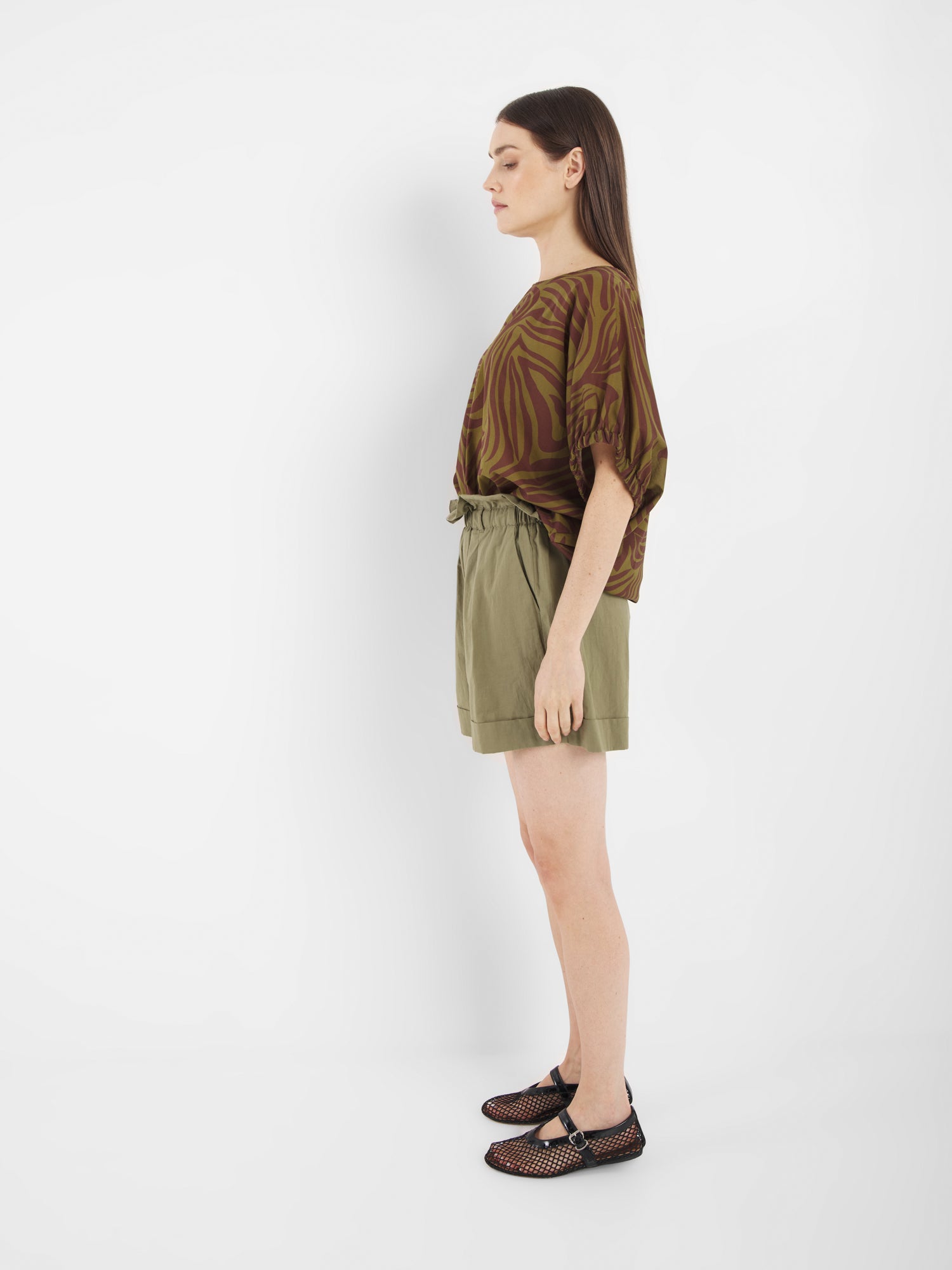 Frau im Profil vor weißem Hintergrund. Sie trägt die Bluse Cowboy Zebra Olive, hochtaillierte, hellolivfarbene Shorts und schwarze Sandalen mit Cut-Out-Design.