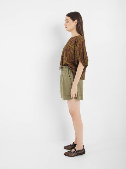 Frau im Profil vor weißem Hintergrund. Sie trägt die Bluse Cowboy Zebra Olive, hochtaillierte, hellolivfarbene Shorts und schwarze Sandalen mit Cut-Out-Design.