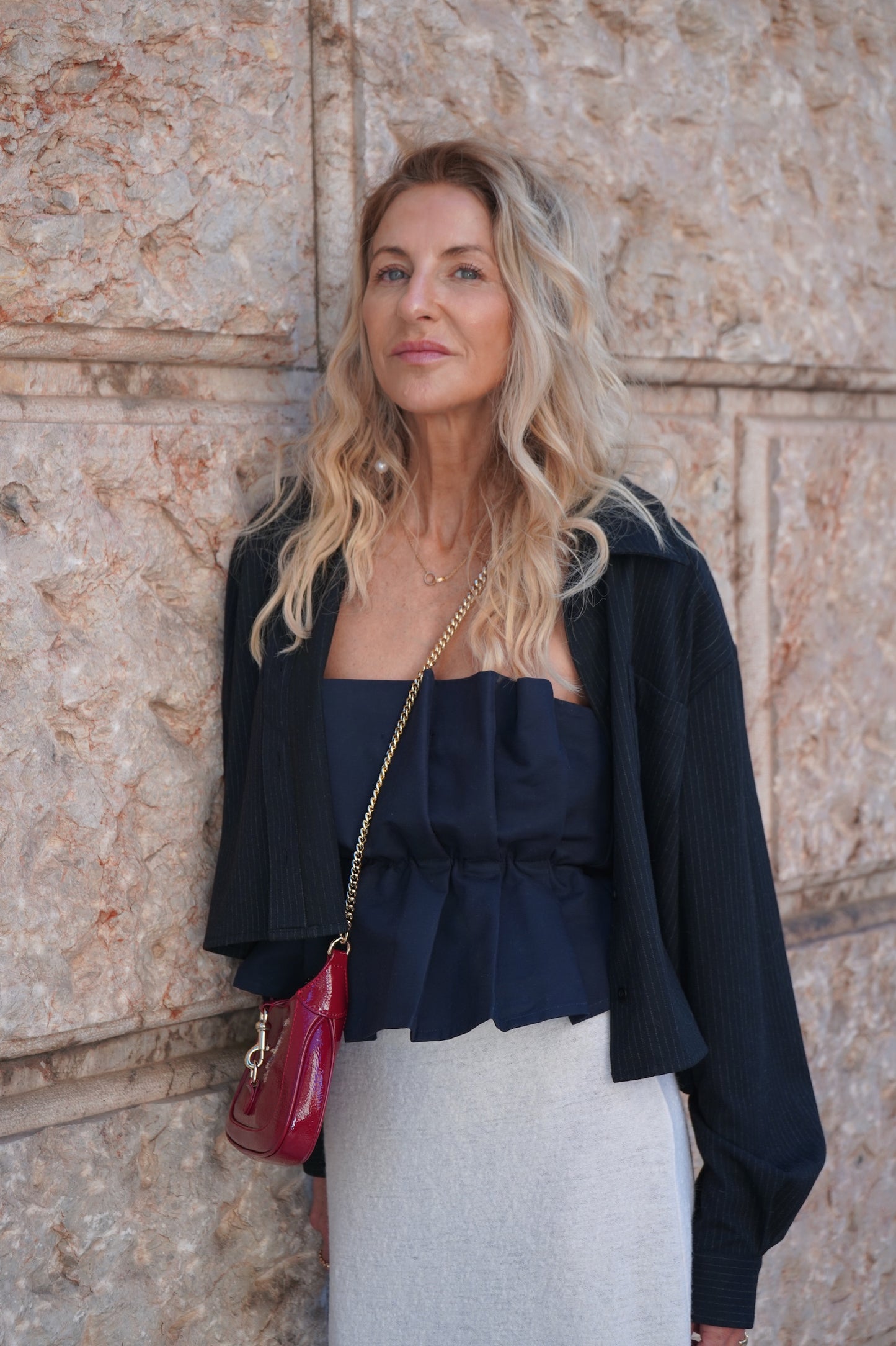 Eine Frau mit langen, gewellten blonden Haaren steht an einer Steinmauer. Sie trägt ein Pleat Top Navy, einen hellgrauen Faltenrock, eine schwarze Jacke und hat eine rote Umhängetasche dabei.