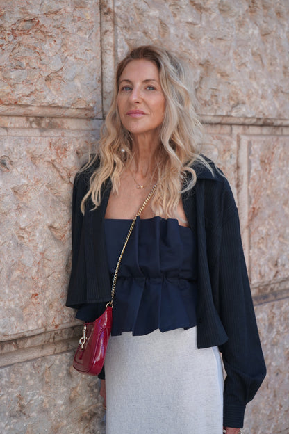 Eine Frau mit langen, gewellten blonden Haaren steht an einer Steinmauer. Sie trägt ein Pleat Top Navy, einen hellgrauen Faltenrock, eine schwarze Jacke und hat eine rote Umhängetasche dabei.
