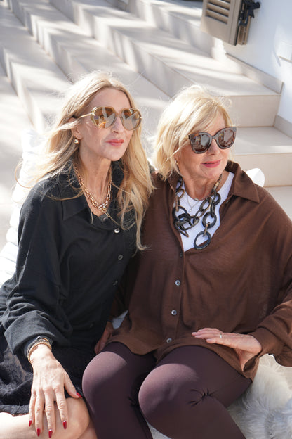 Zwei ältere Frauen mit blonden Haaren, Sonnenbrillen und stilvollen Outfits - darunter die Bluse Antonia Cashmere Black One Size von Ella Silla x SOSUE - sitzen dicht beieinander auf einer Außentreppe, lächeln und genießen den sonnigen Tag.