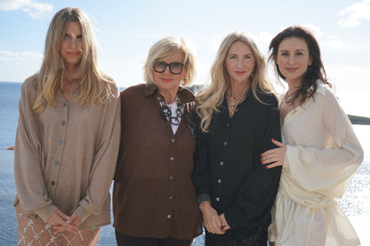 Vier Frauen stehen gemeinsam im Freien am Wasser und lächeln in die Kamera. Eine trägt die stilvolle Bluse Antonia Cashmere Black One Size von Ella Silla x SOSUE und strahlt luxuriöse Eleganz aus, während hinter ihnen das Sonnenlicht scheint.