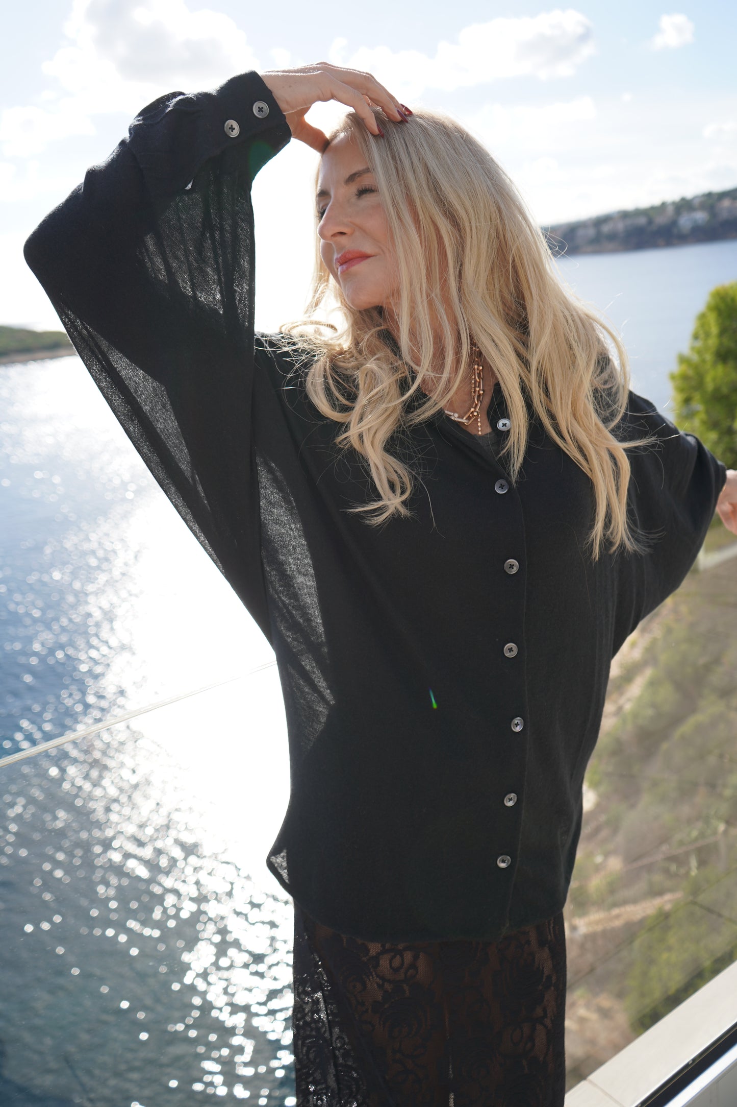 Eine Frau mit langen blonden Haaren posiert anmutig auf einem Balkon mit Blick auf das Wasser. Sie trägt die Bluse Antonia Cashmere Black One Size von Ella Silla x SOSUE, während das Sonnenlicht auf der Oberfläche schimmert und das Grün im Hintergrund zu sehen ist.
