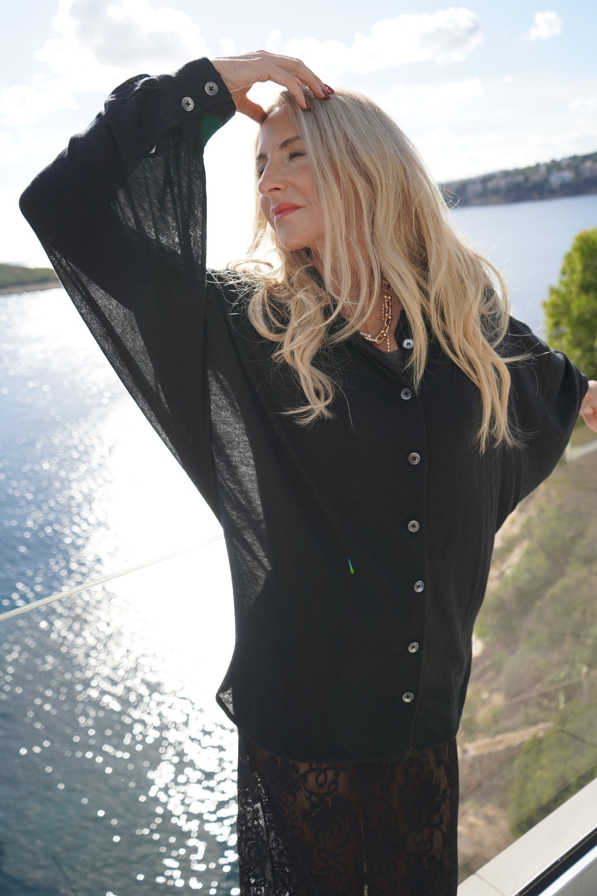 Eine Frau mit langen blonden Haaren posiert anmutig auf einem Balkon mit Blick auf das Wasser. Sie trägt die Bluse Antonia Cashmere Black One Size von Ella Silla x SOSUE, während das Sonnenlicht auf der Oberfläche schimmert und das Grün im Hintergrund zu sehen ist.