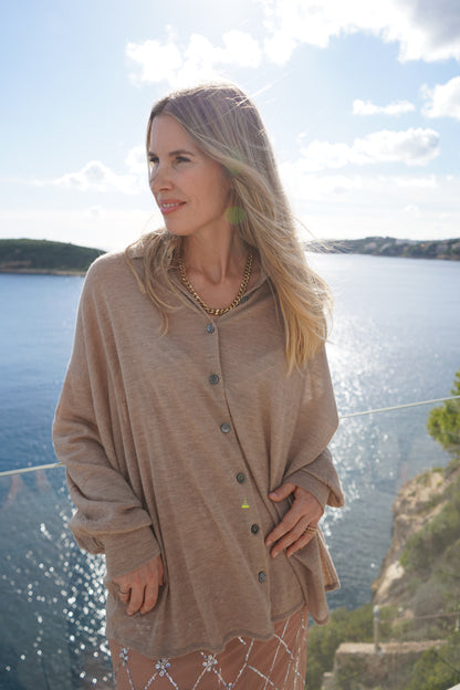 Eine Frau mit langen blonden Haaren, die die Bluse Antonia Cashmere Taupe One Size Strickjacke von Ella Silla x SOSUE und eine goldene Halskette trägt, steht im Freien in der Nähe von Wasser und fernen Hügeln unter einem teilweise bewölkten Himmel.