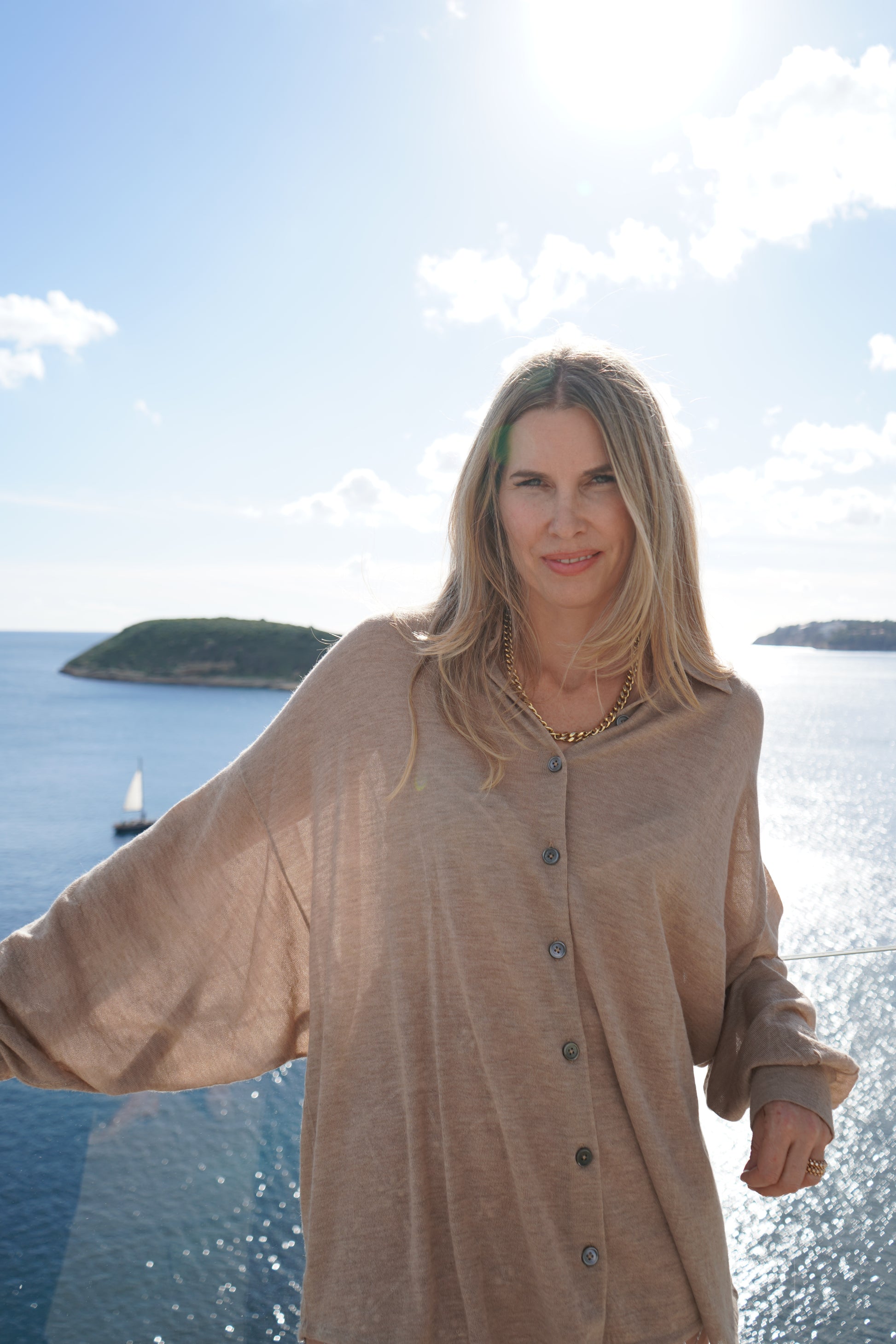 Eine Frau mit langen blonden Haaren steht am Meer und trägt die Bluse Antonia Cashmere Taupe One Size von Ella Silla x SOSUE und eine goldene Halskette. Über ihr scheint die Sonne, im Hintergrund sind eine Insel und ein Segelboot zu sehen.