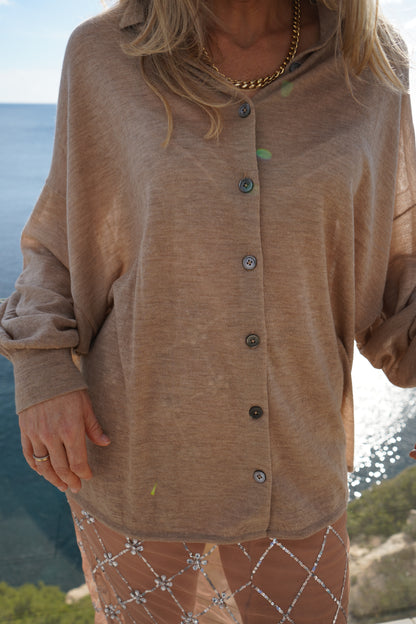 Eine Person, die die Bluse Antonia Cashmere Taupe One Size von Ella Silla x SOSUE und eine goldene Halskette trägt, steht im Freien in der Nähe von Wasser, im Sonnenlicht und mit Bäumen im Hintergrund. Nur der Oberkörper ist sichtbar und strahlt luxuriöse Eleganz aus.