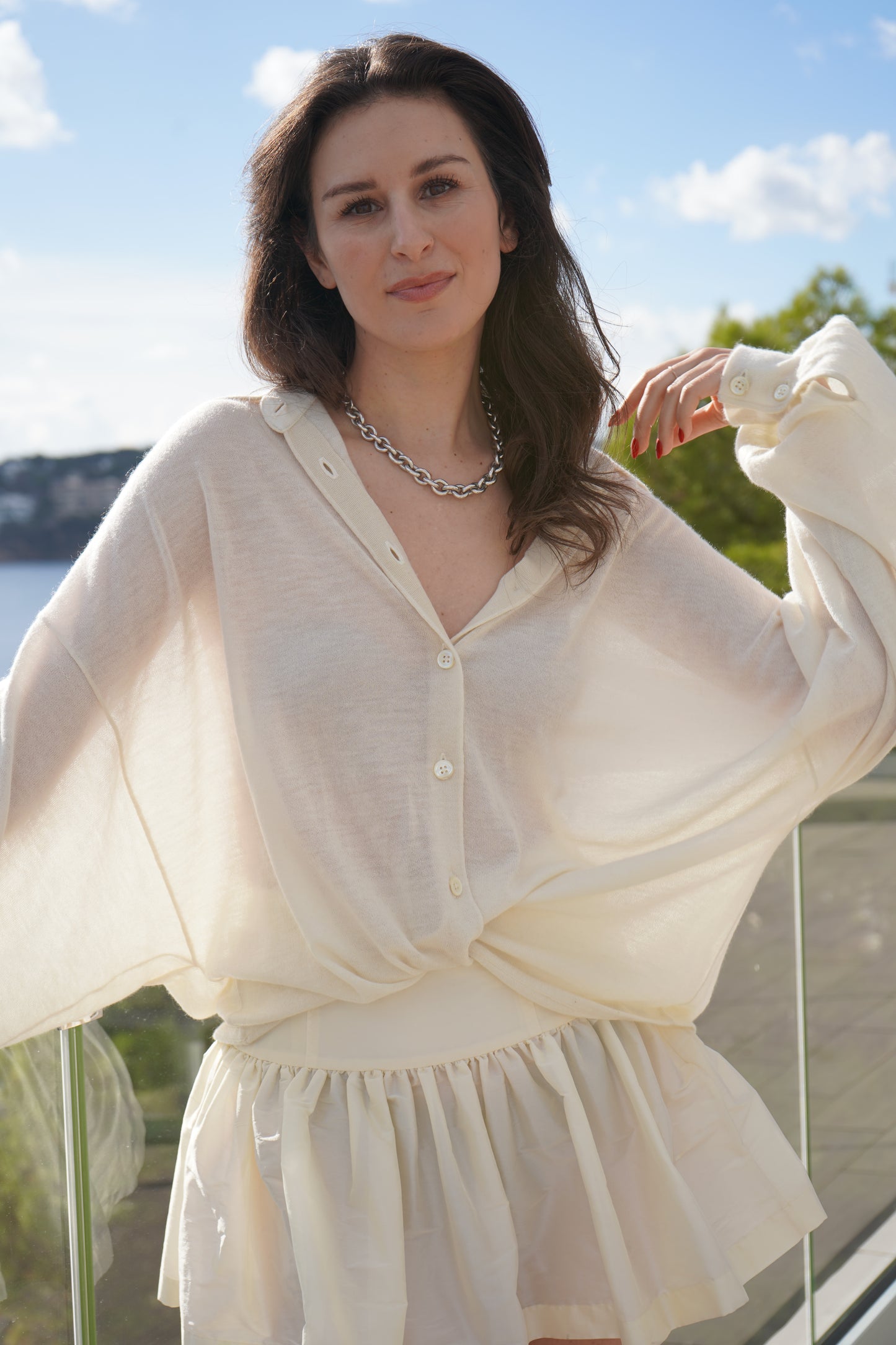 Eine Frau mit langen braunen Haaren, die die Bluse Antonia Cashmere Ecru One Size von Ella Silla x SOSUE und einen dazu passenden Rock trägt, steht im Freien an einem Glasgeländer und lächelt sanft mit dem Grün und dem blauen Himmel im Hintergrund.
