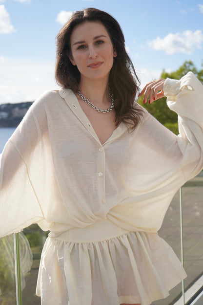Eine Frau mit langen braunen Haaren, die die Bluse Antonia Cashmere Ecru One Size von Ella Silla x SOSUE und einen dazu passenden Rock trägt, steht im Freien an einem Glasgeländer und lächelt sanft mit dem Grün und dem blauen Himmel im Hintergrund.