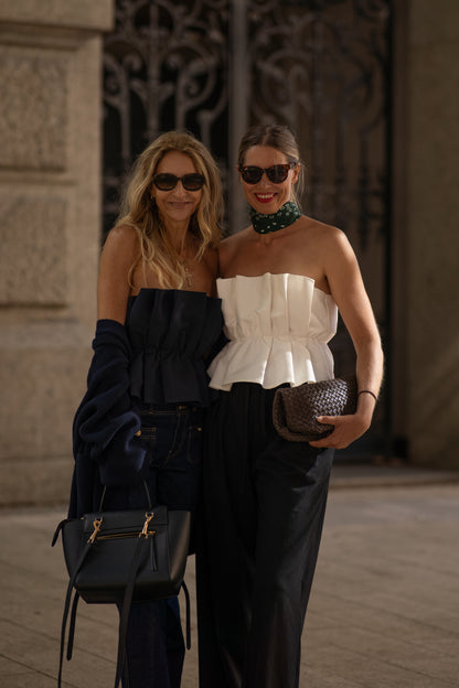 Zwei Frauen stehen dicht beieinander und lächeln mit Sonnenbrillen. Die eine trägt das Pleat Top Navy und eine dunkle Hose, in der Hand eine schwarze Tasche. Die andere kombiniert ihr weißes trägerloses Oberteil und die dunkle Hose mit einem grünen Schal und einer gewebten Clutch.