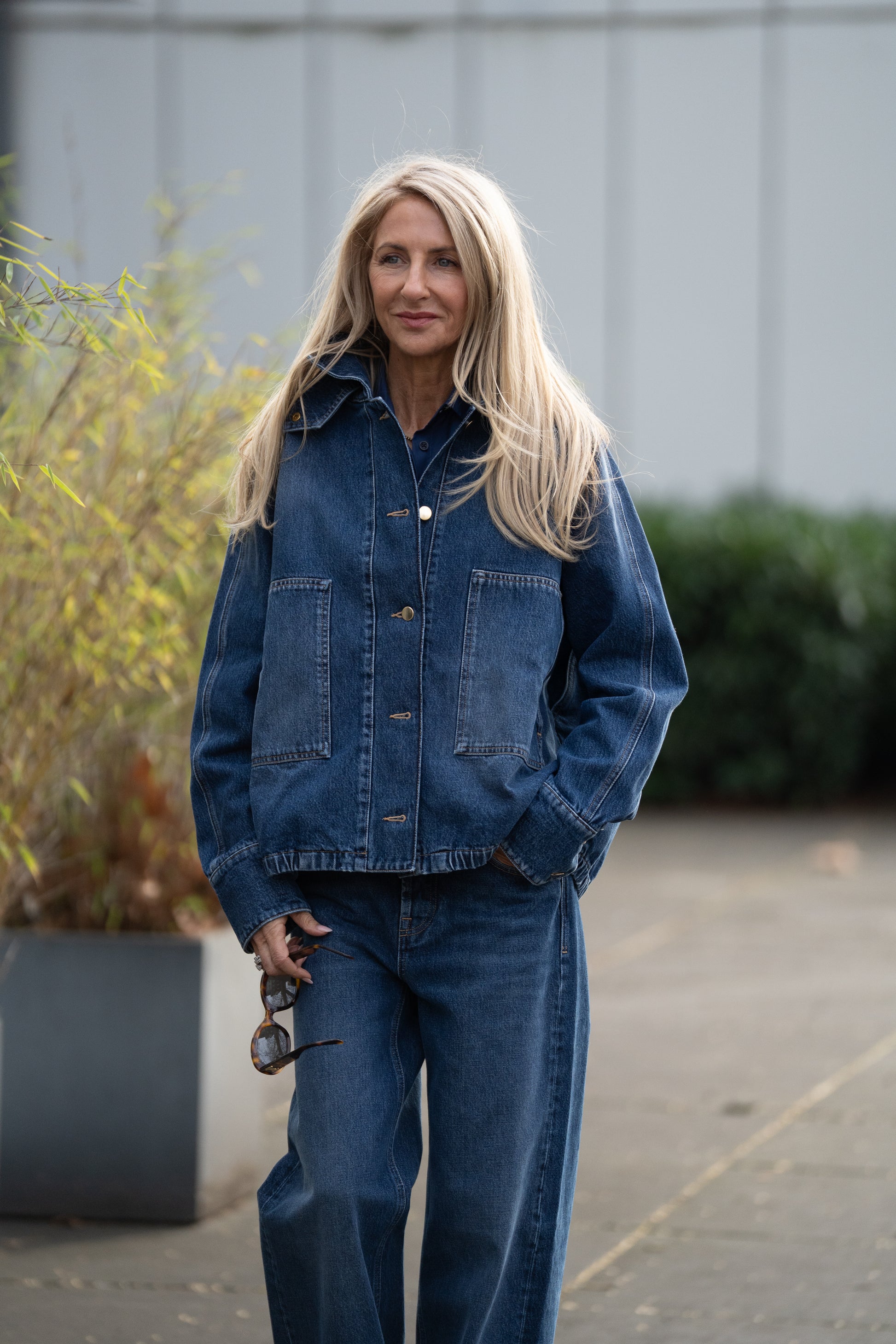 Eine Frau mit langen blonden Haaren steht im Freien in einer locker sitzenden Bomberjacke und einer dazu passenden Jeans mit weitem Bein, hält eine Brille in der Hand und lächelt sanft, während im Hintergrund Grünflächen und ein modernes Gebäude zu sehen sind.