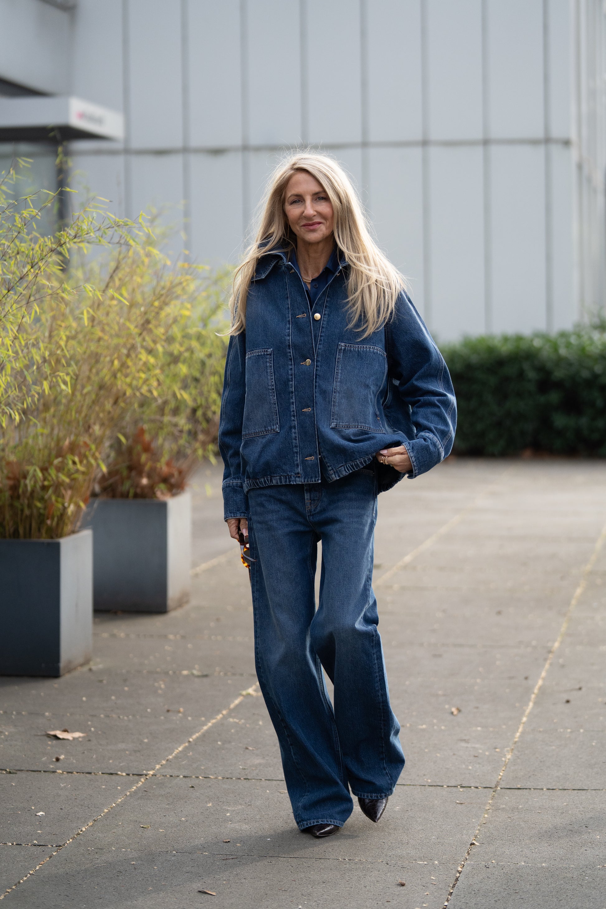 Eine Frau mit langen blonden Haaren trägt die Jacke Bomber Denim und Jeans mit weitem Bein. Sie steht im Freien auf einer gepflasterten Fläche in der Nähe großer Pflanzentöpfe und eines modernen Gebäudes.
