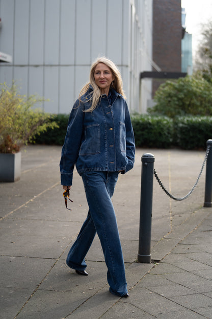 Eine Frau mit langen blonden Haaren geht draußen auf einem Bürgersteig spazieren. Sie trägt die Jacke Bomber Denim und eine Denim-Jeans. Sie trägt eine Sonnenbrille, lächelt leicht und ist umgeben von Grün und einem modernen Gebäude im Hintergrund.
