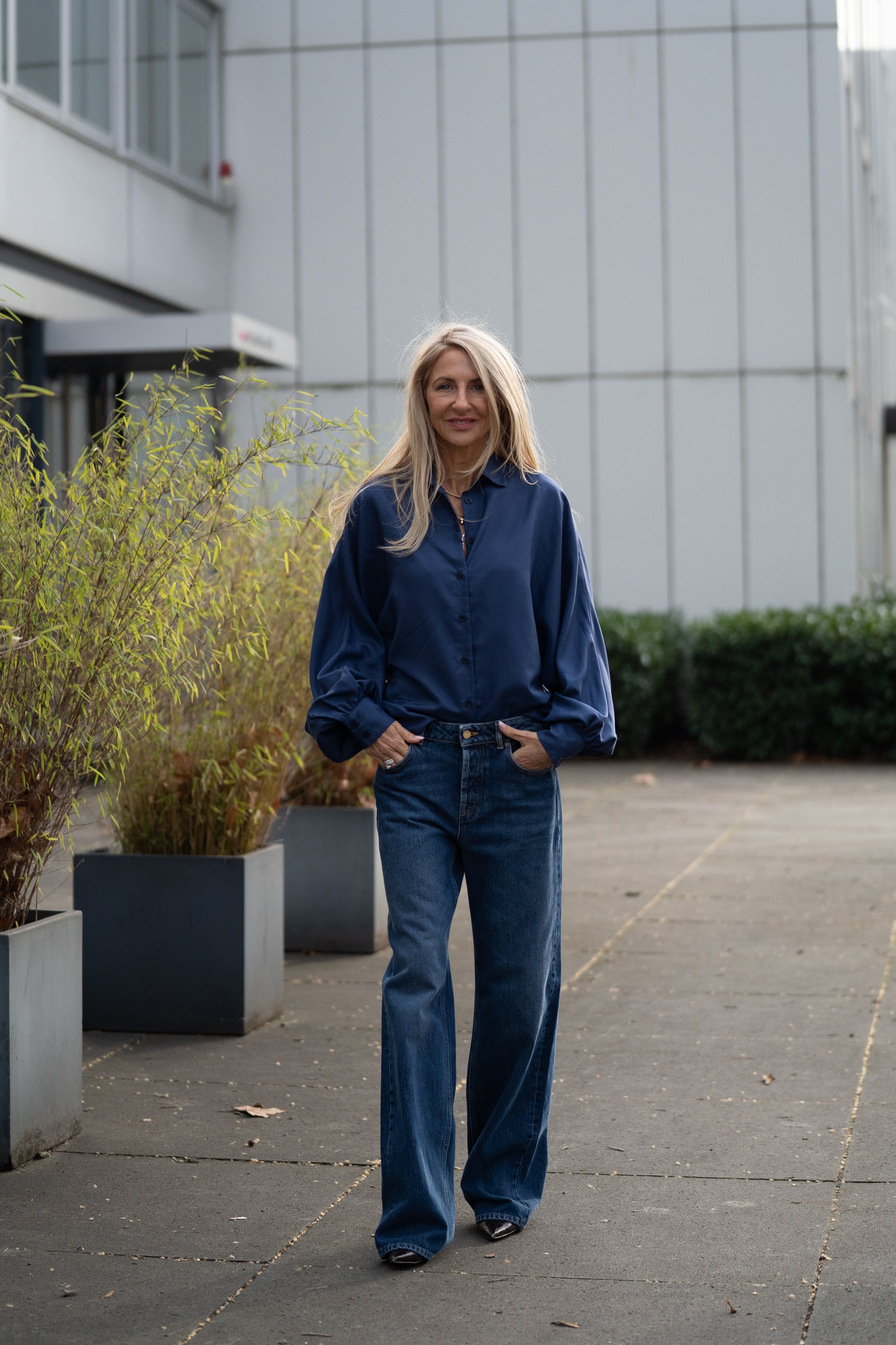 Eine Frau mit langen blonden Haaren, die eine blaue Bluse und eine „NOLA Jeans Dark Blue – Wide Leg Damenjeans aus Premium-Denim, Made in Italy“ trägt, steht im Freien auf einem gepflasterten Gehweg zwischen großen Pflanzkübeln und modernen Gebäuden.