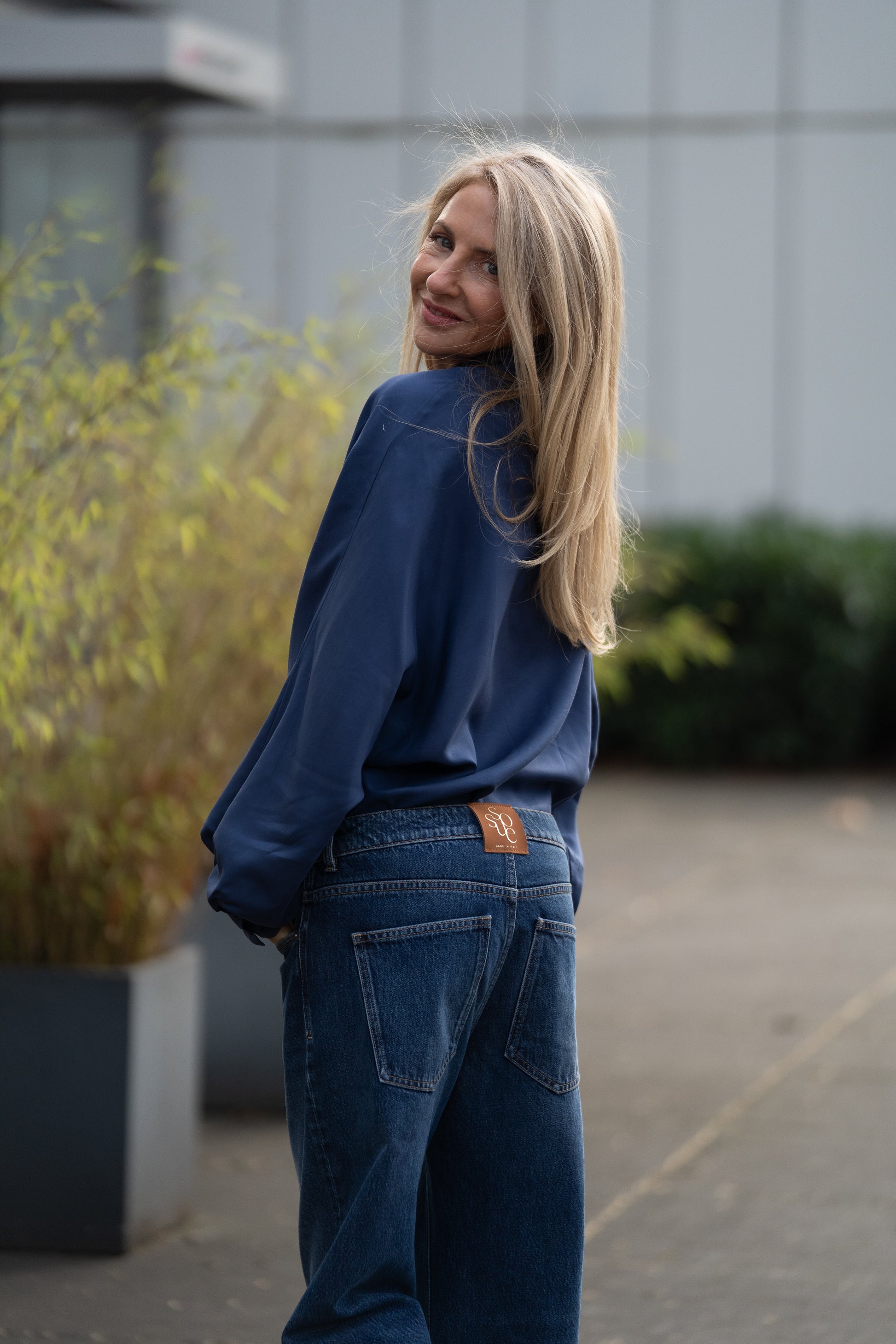 Eine Frau mit langen blonden Haaren, die ein lockeres blaues Shirt und eine „NOLA Jeans Light Blue – Wide Leg Damenjeans aus Premium-Denim, Made in Italy“ trägt, steht lächelnd auf einem Bürgersteig, im städtischen Hintergrund sind grüne Pflanzen und ein graues Gebäude zu sehen.