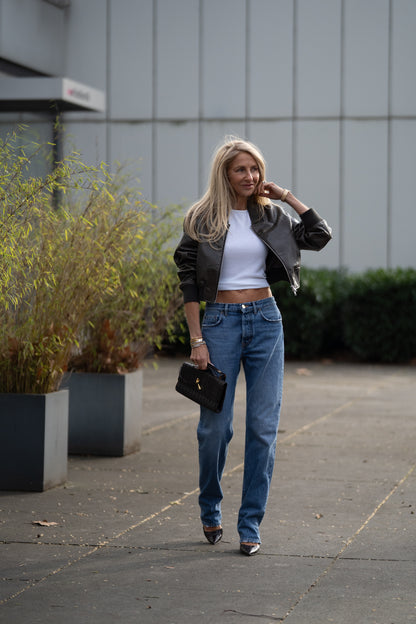 Eine Frau mit langen blonden Haaren trägt eine „SIENNA Jeans Light Blue – Straight Leg Damenjeans aus Premium-Denim, Made in Italy“, ein kurzes weißes Oberteil, eine schwarze Jacke und Pumps, während sie mit einer schwarzen Handtasche im Freien an Blumentöpfen und einem modernen Gebäude vorbeigeht.