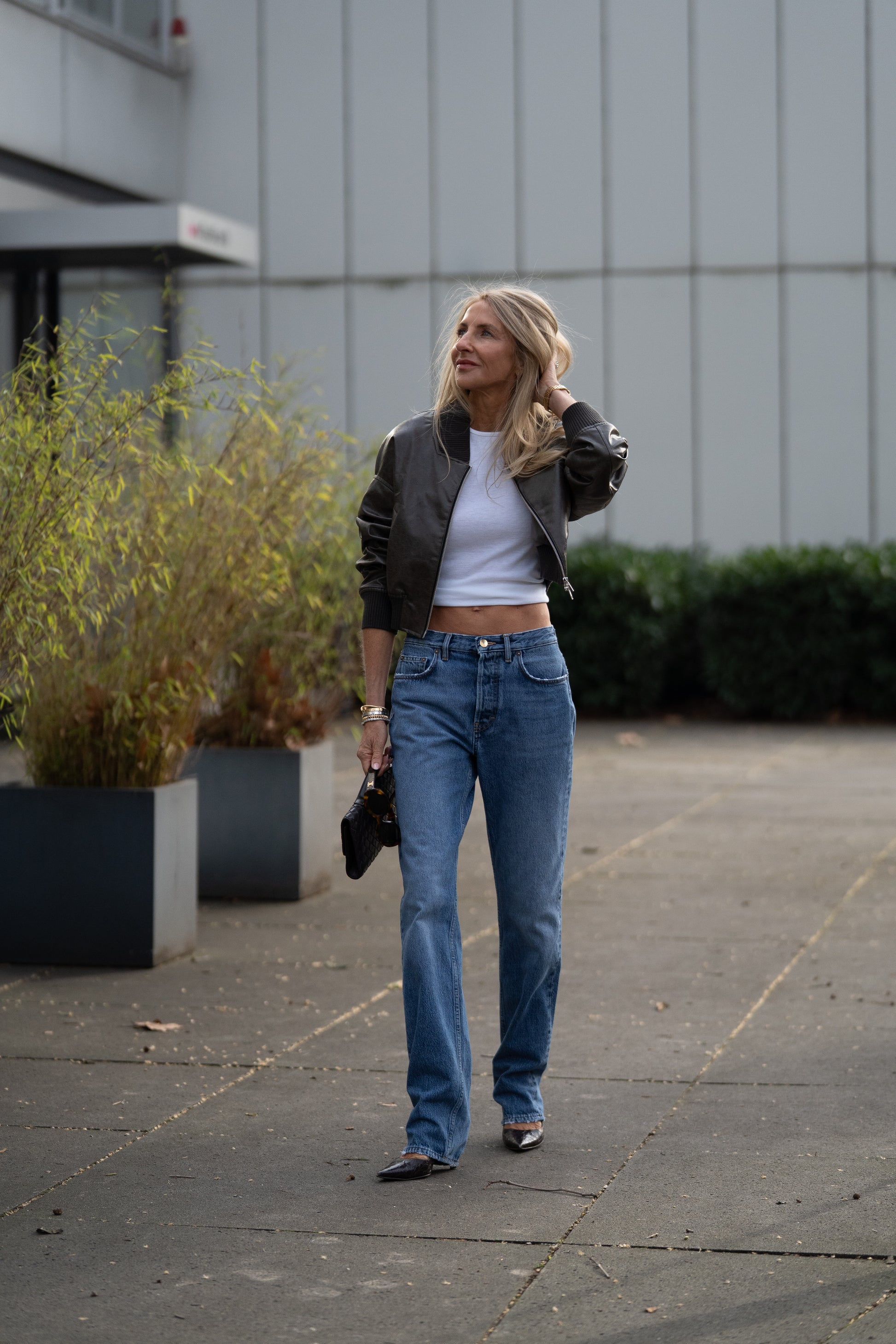 Eine Frau mit langen blonden Haaren geht draußen in hellblauen SIENNA-Jeans spazieren – Straight-Leg-Damenjeans aus Premium-Denim, hergestellt in Italien –, kombiniert mit einer grauen Bomberjacke, einem weißen Crop-Top, spitz zulaufenden Schuhen und einer kleinen schwarzen Handtasche.