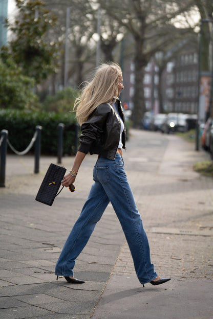 Eine Frau mit langen blonden Haaren geht selbstbewusst auf einem Gehweg, trägt Jeans Straight Sienna Dark Blue, schwarze Absätze, eine schwarze Kurzjacke und eine schwarze Handtasche. Bäume und Gebäude verschwimmen im Hintergrund.