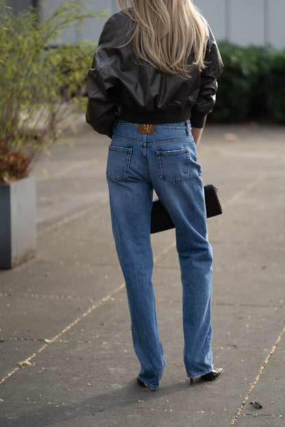 Eine Person mit langen blonden Haaren, die eine schwarze Lederjacke und eine „SIENNA Jeans Light Blue – Straight Leg Damenjeans aus Premium-Denim, Made in Italy“ trägt, hält eine schwarze Clutch in der Hand und steht im Freien auf dem Bürgersteig in der Nähe von Grünflächen, mit dem Rücken zur Kamera.