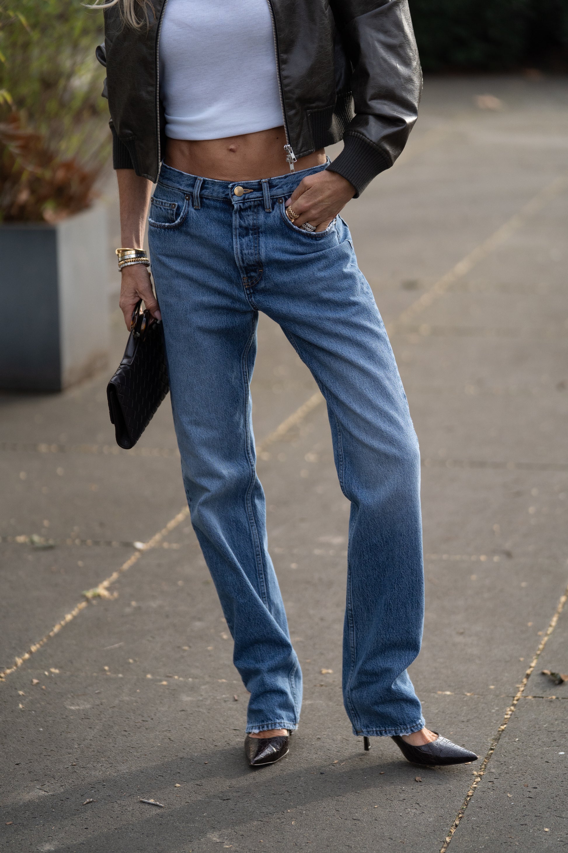 Eine Person steht im Freien und trägt eine „SIENNA Jeans Light Blue – Straight Leg Damenjeans aus Premium-Denim, Made in Italy“, ein weißes Crop-Top, eine schwarze Lederjacke, schwarze spitze Schuhe und hält eine schwarze Clutch in der Hand. Ihr Gesicht ist nicht zu sehen.