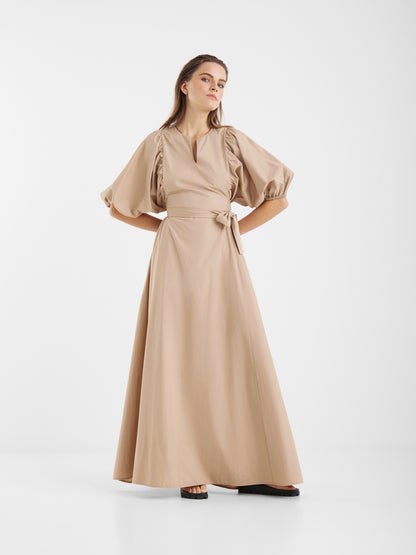 Eine Frau steht selbstbewusst in dem Kleid Antonia Lang, einem beigen Maxikleid mit gepufften Halbärmeln, taillierter Passform mit Bindegürtel und ausgestelltem Rock, das vor einem schlichten weißen Hintergrund feminine Eleganz ausstrahlt.