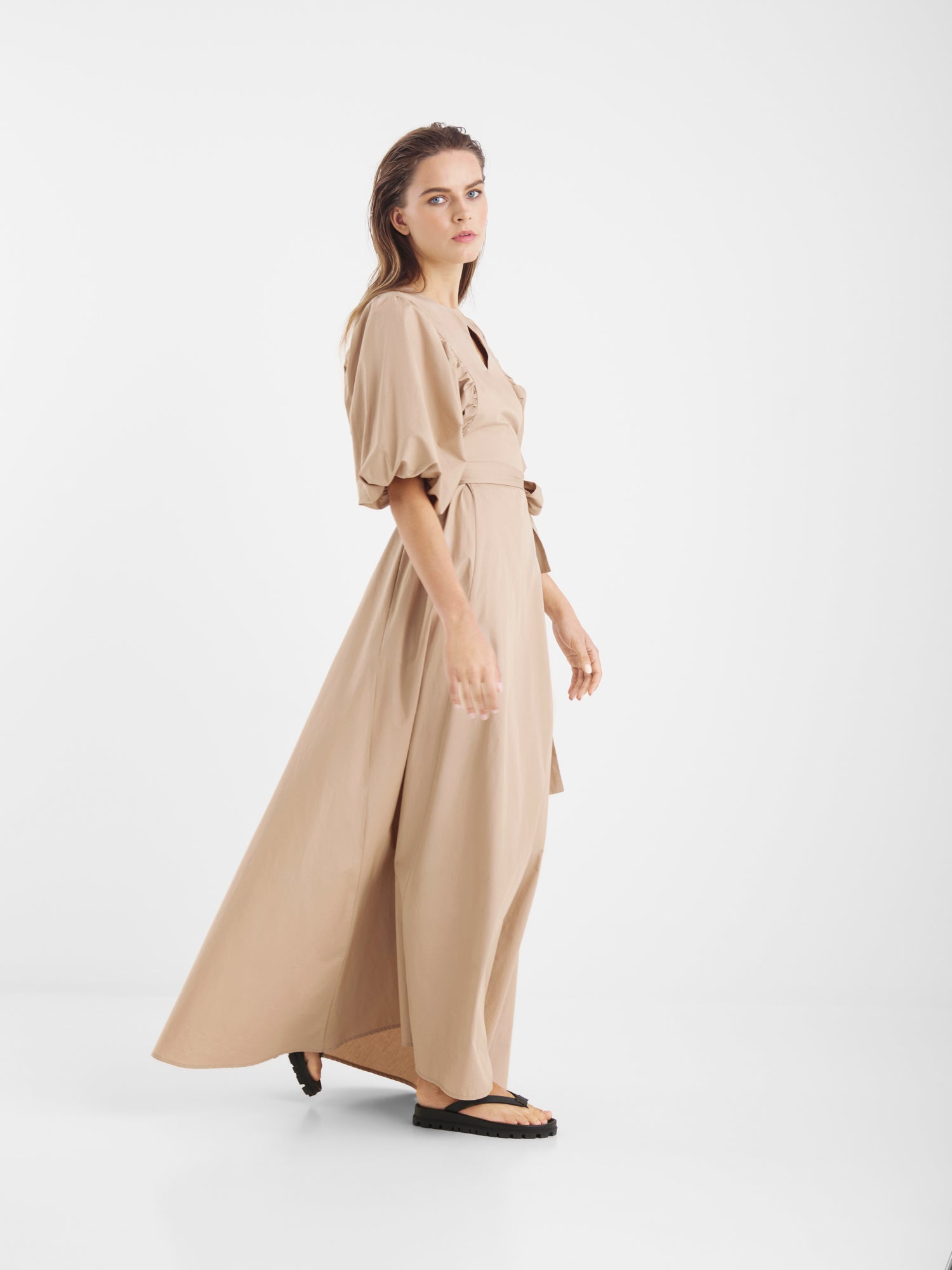 Eine Frau steht im Profil vor einem weißen Hintergrund und trägt das Kleid Antonia Lang - ein beigefarbenes Maxikleid mit kurzen Puffärmeln und einem fließenden Rock -, dazu schwarze flache Sandalen und offenes, schulterlanges Haar.