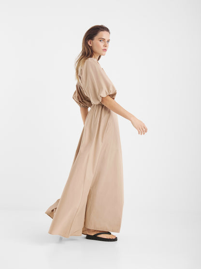 Eine Frau im Kleid Antonia Lang, einem beigen Maxikleid und schwarzen Sandalen steht im Profil vor einem weißen Hintergrund. Ihr locker frisiertes Haar und ihr gleichmäßiger Schritt verkörpern feminine Eleganz, während sie über ihre Schulter blickt.