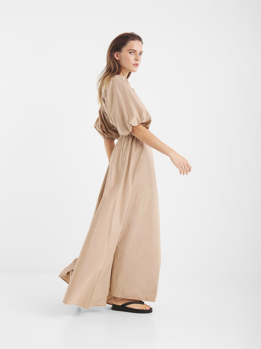 Die Frau trägt das lockere hellbraune Maxikleid Hey Alice ! und schwarze Sandalen. Sie steht im Profil vor einem schlichten weißen Hintergrund und blickt über ihre Schulter, während sie vorwärts geht.