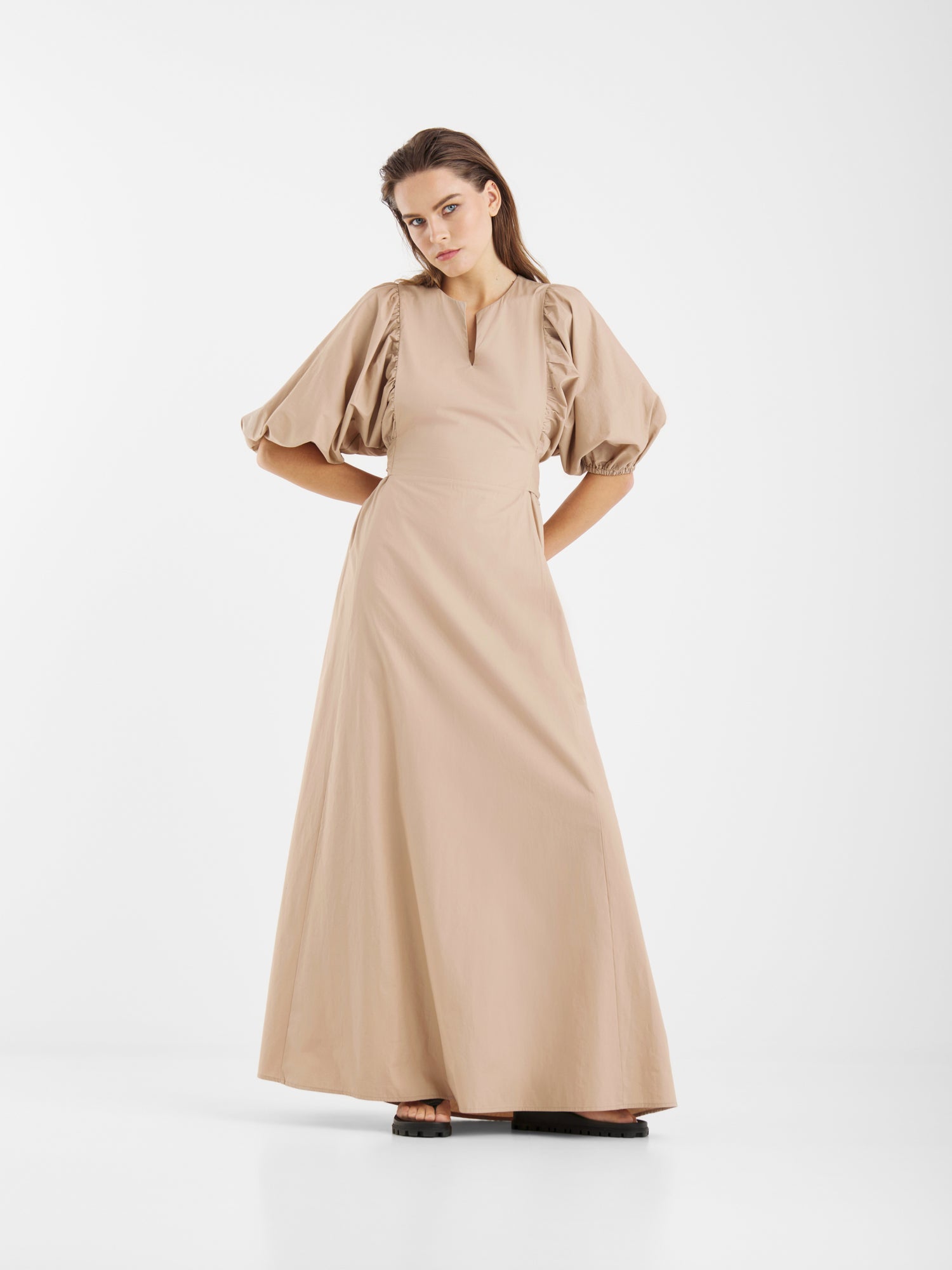 Eine Frau posiert vor einem weißen Hintergrund und trägt das Kleid Antonia Lang, ein beiges Maxikleid mit Puffärmeln und Schlüssellochausschnitt. Sie strahlt feminine Eleganz aus, die Hände in die Hüften gestemmt und den Blick leicht nach unten gerichtet.