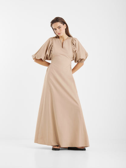 Eine Frau posiert vor einem weißen Hintergrund und trägt das Kleid Antonia Lang, ein beiges Maxikleid mit Puffärmeln und Schlüssellochausschnitt. Sie strahlt feminine Eleganz aus, die Hände in die Hüften gestemmt und den Blick leicht nach unten gerichtet.