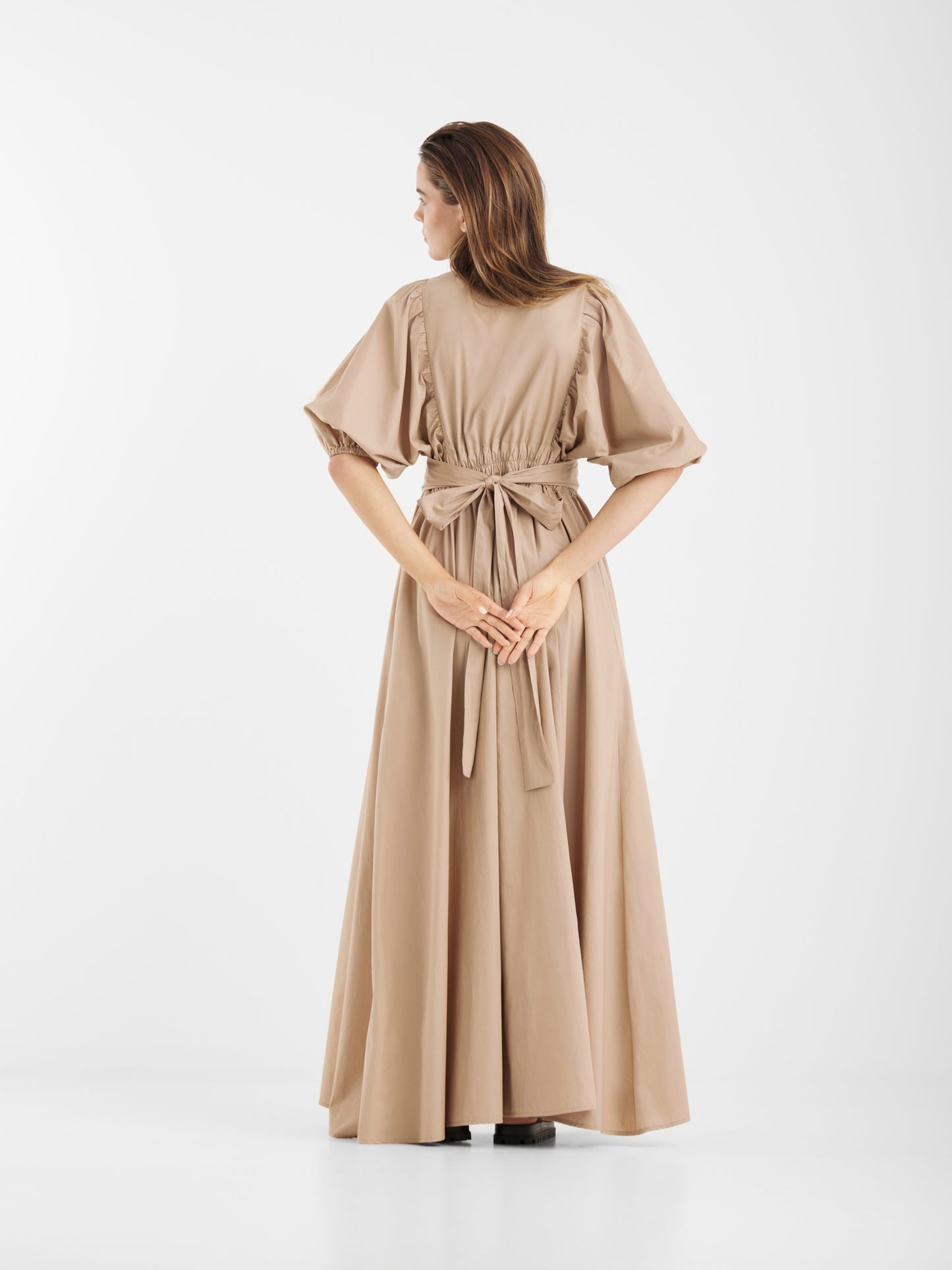 Eine Frau in dem beigen Kleid Antonia Lang steht mit dem Rücken zur Kamera, die Hände verschränkt. Das Kleid hat kurze Ärmel, eine gebundene Taille und einen fließenden Rock, alles vor einem schlichten weißen Hintergrund.