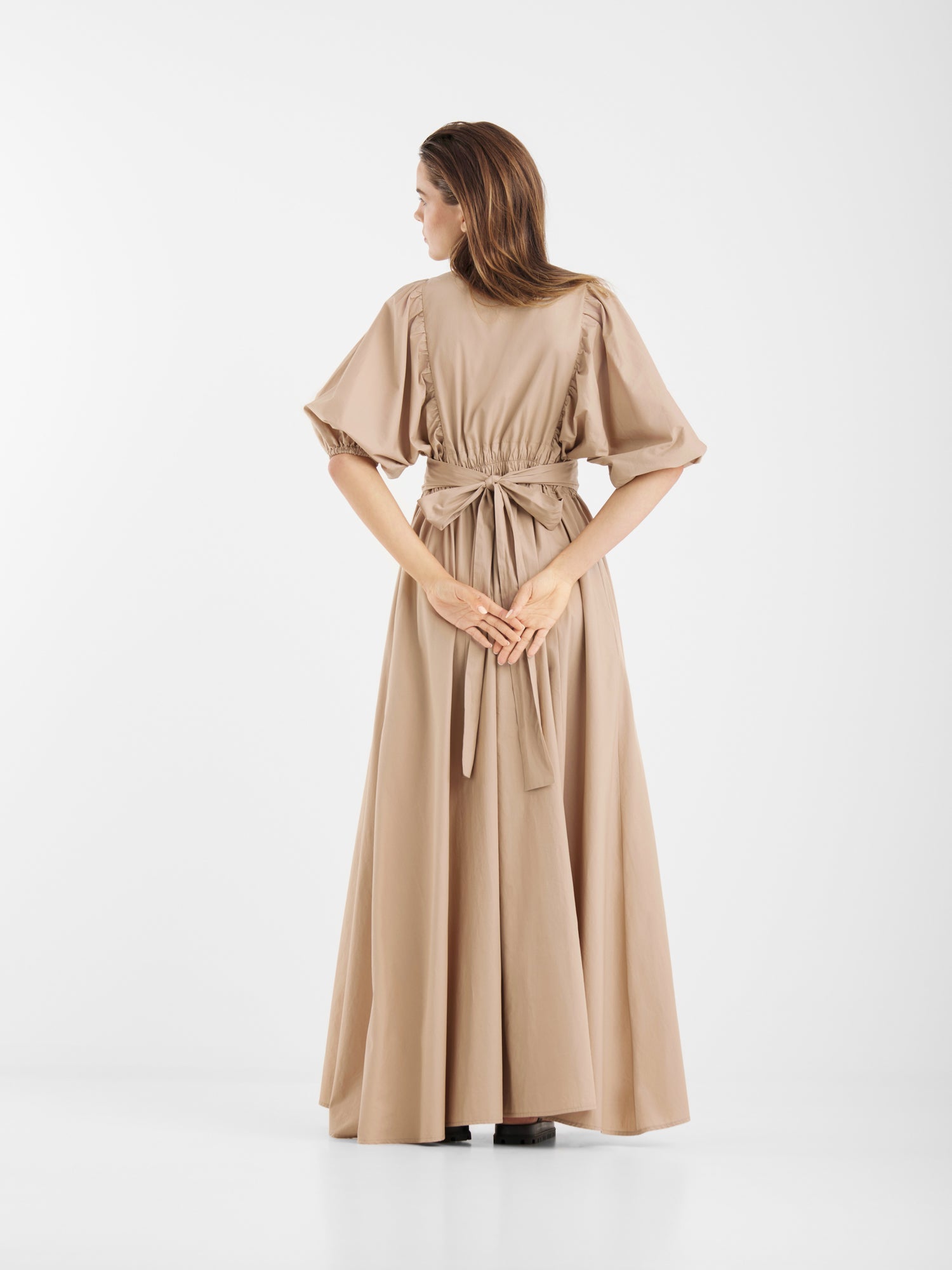 Eine Frau in dem beigen Kleid Antonia Lang steht mit dem Rücken zur Kamera, die Hände verschränkt. Das Kleid hat kurze Ärmel, eine gebundene Taille und einen fließenden Rock, alles vor einem schlichten weißen Hintergrund.